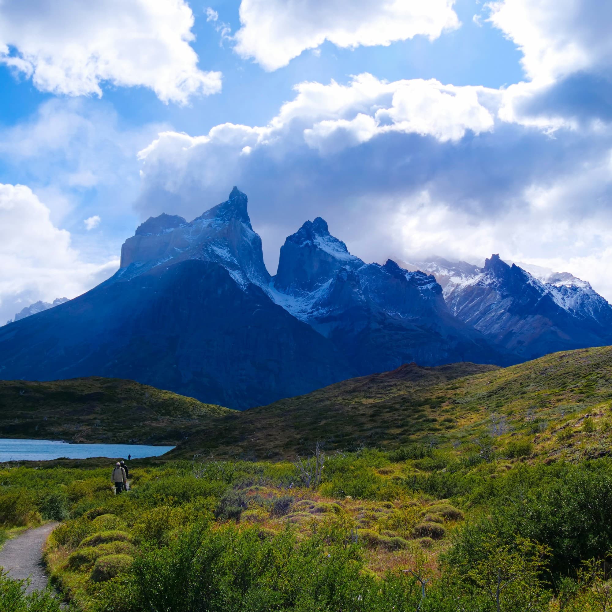 A scenic landscape with towering mountains, a clear sky and a trail leading towards the peaks.