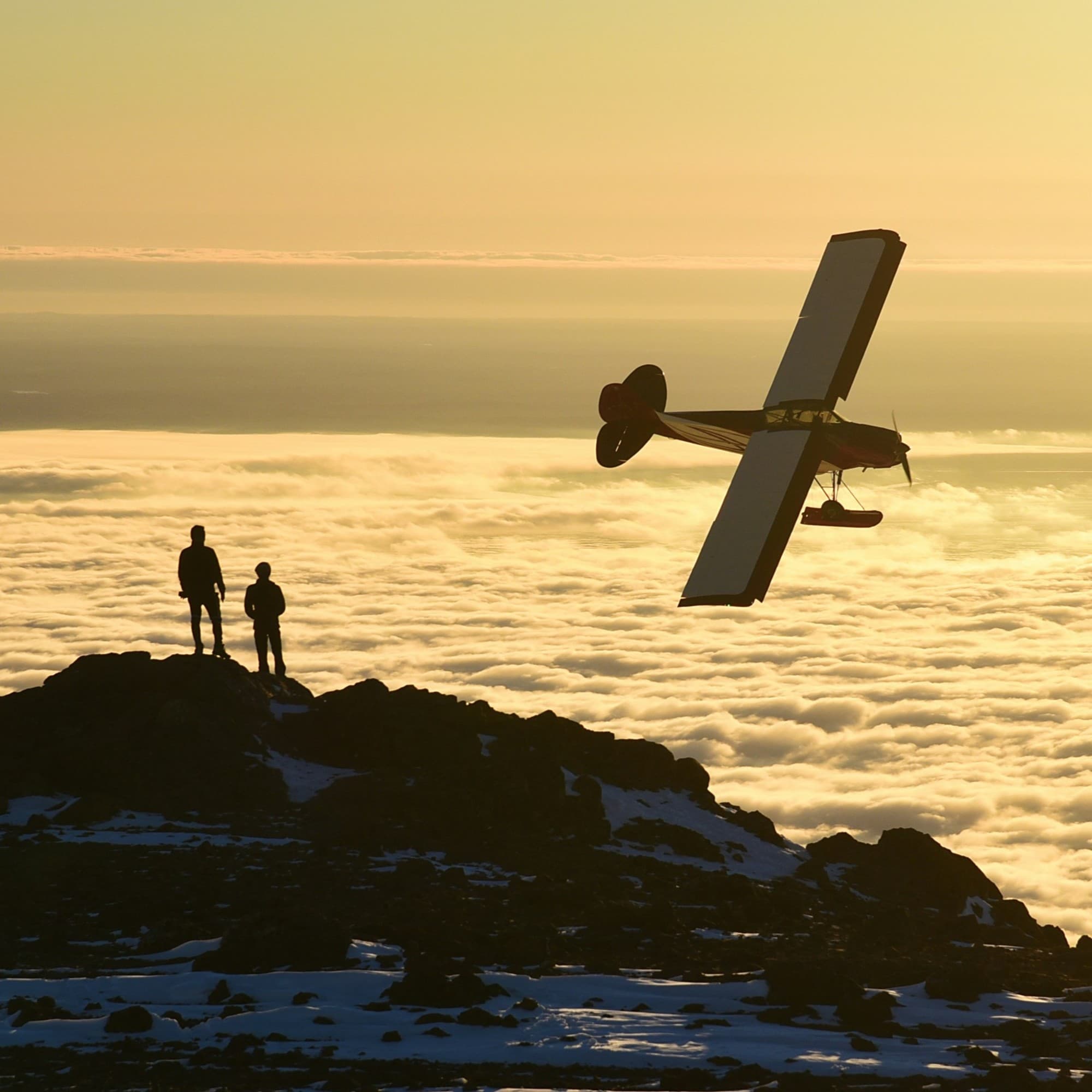 Two people stand on a rocky outcrop above a sea of clouds, with an airplane flying nearby during a stunning sunset or sunrise.