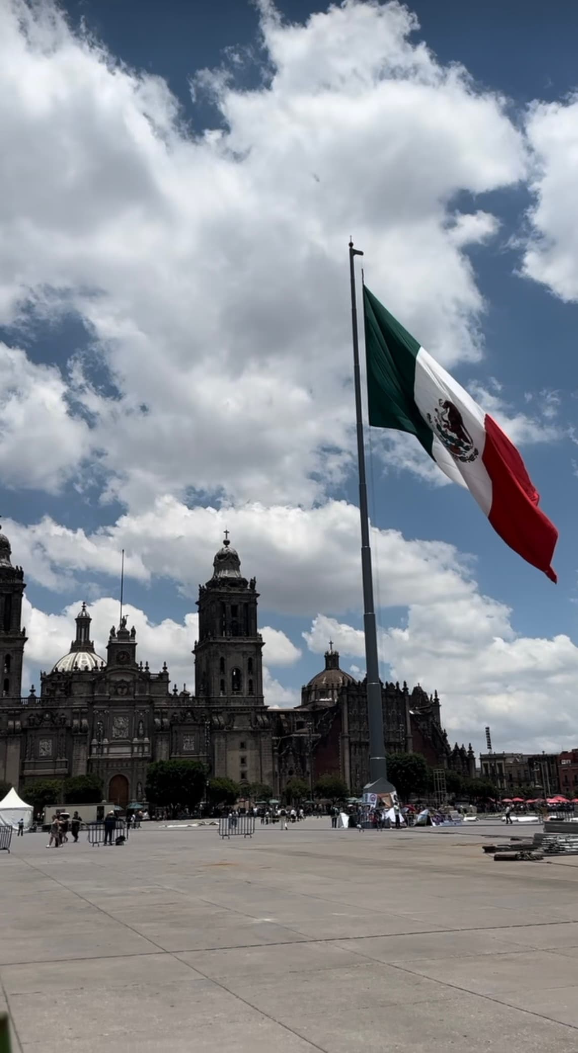 The flag of Mexico City at a plaza with a historic building and a cloudy sky in the background.