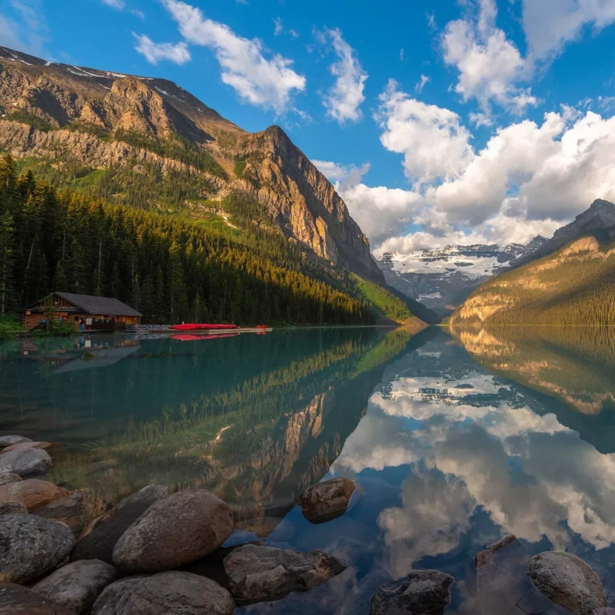 The image shows a serene lake reflecting mountains and clouds, with a cabin and canoes to the left, set in a tranquil forested area.