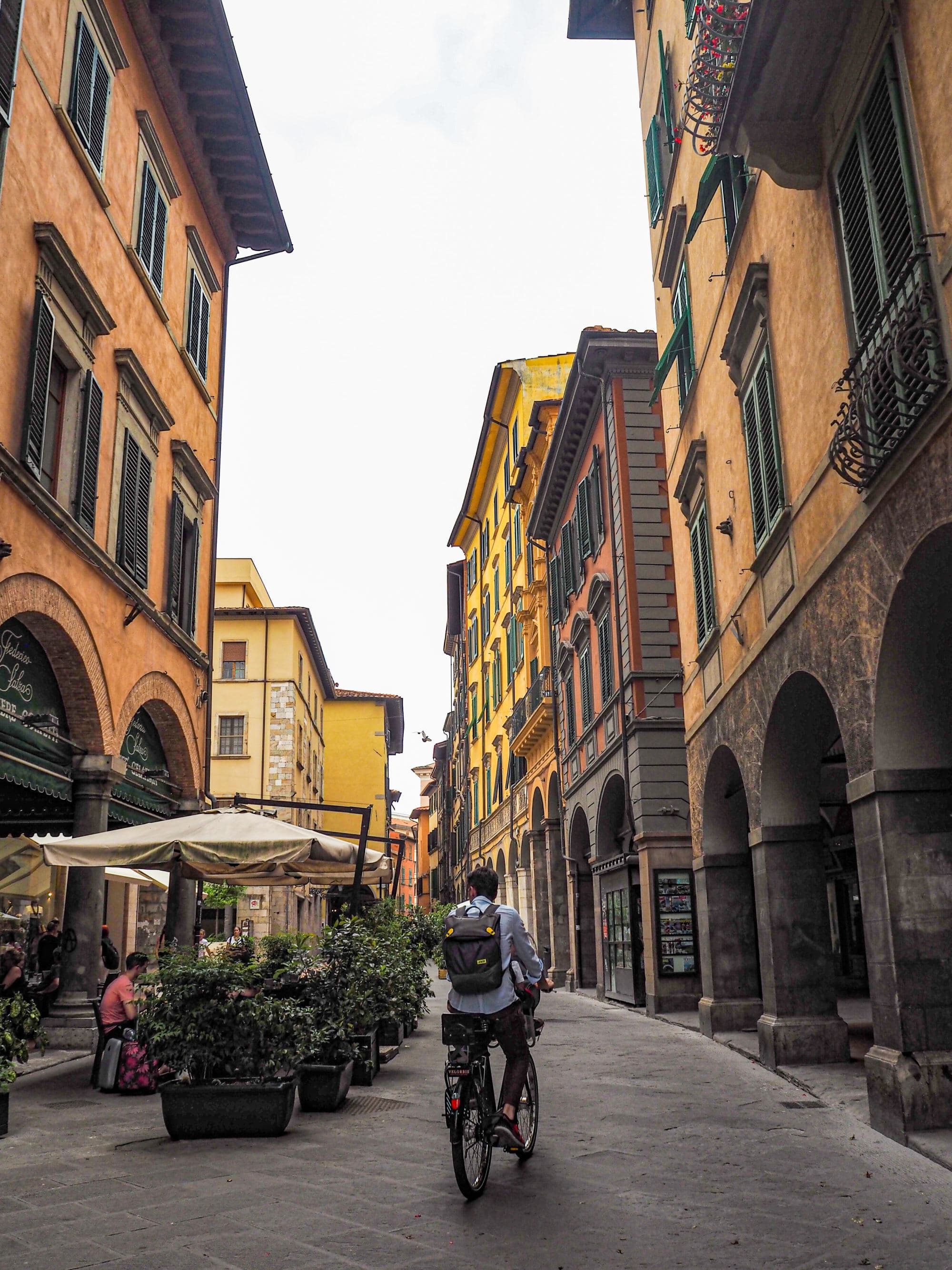 A person on a bicycle and people sitting on side walk in a street with buildings.