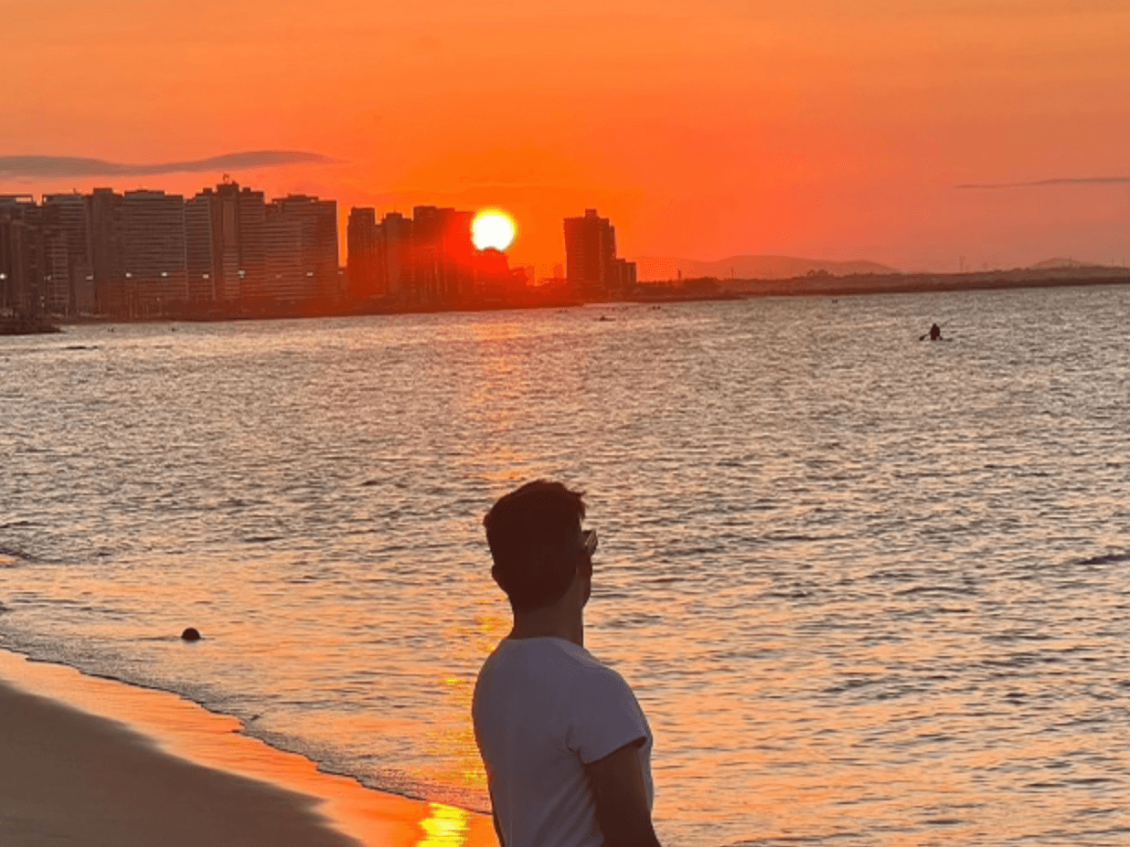 A man looking out to a beaming orange sunset peaking through the city skyline and shining onto the water from the beach.