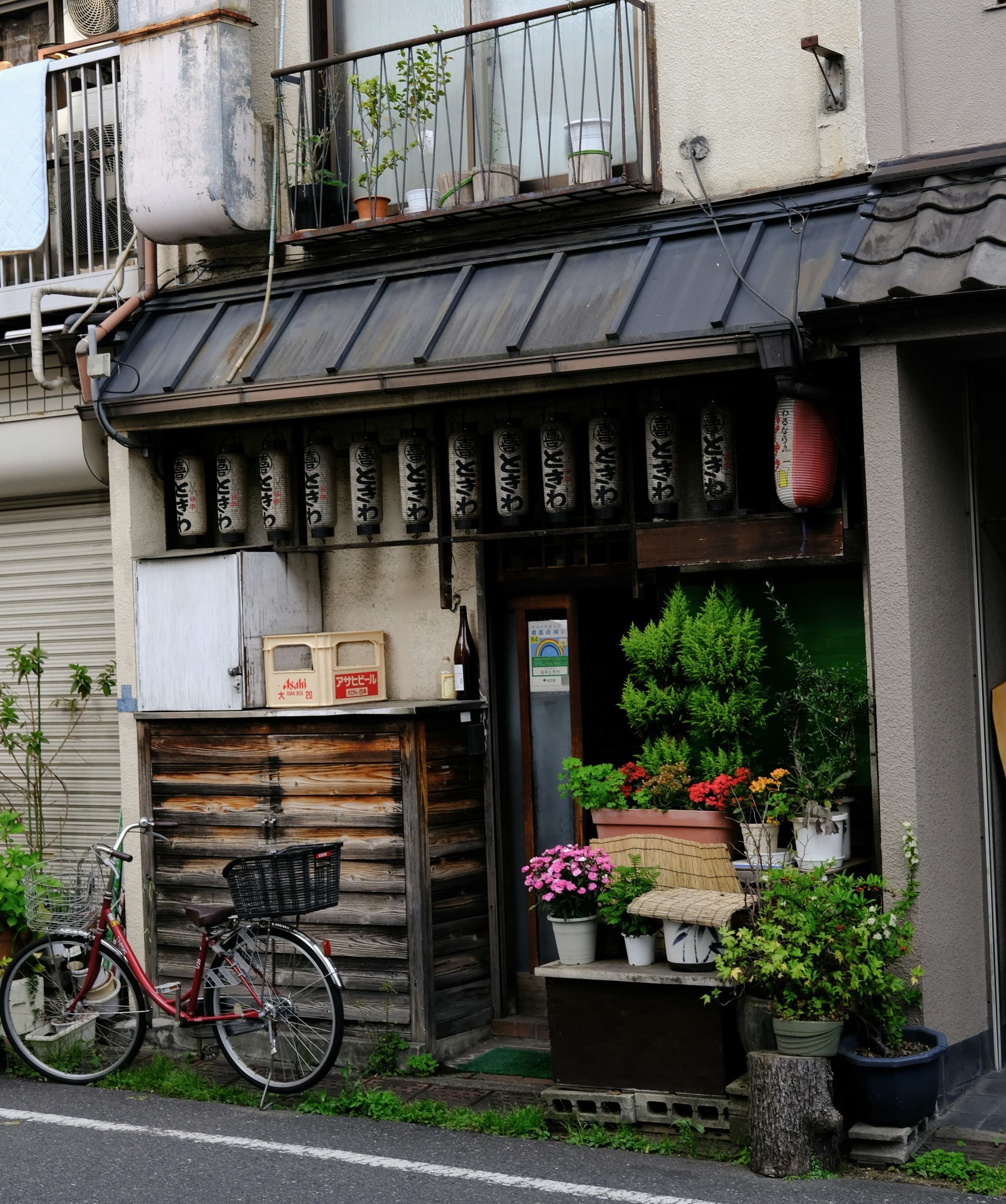 A building with Japanese architecture that showcases hanging lanterns, potted plants and a bicycle parked outside in front of it.