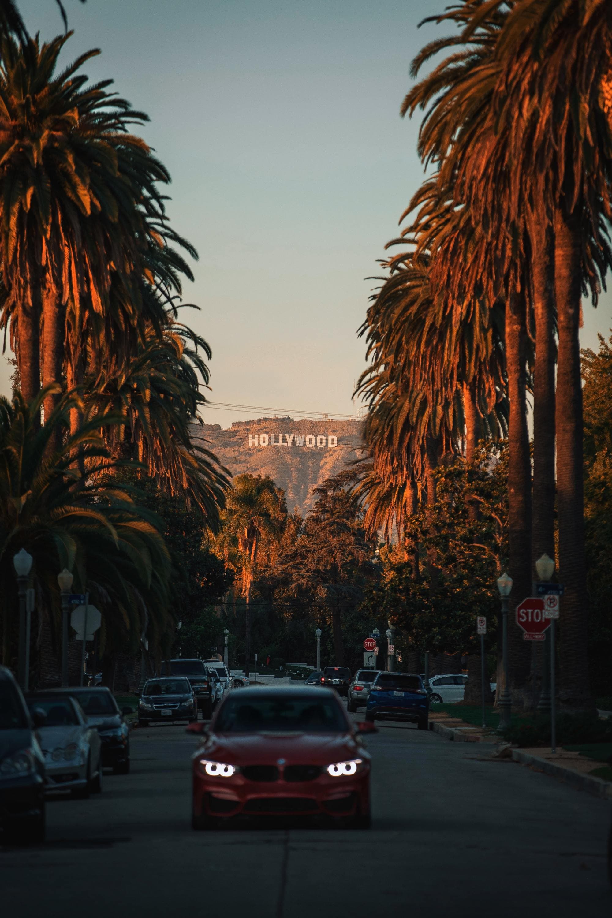 a red car drives down a road lined with palm trees at sunset with the Hollywood sign in the distance