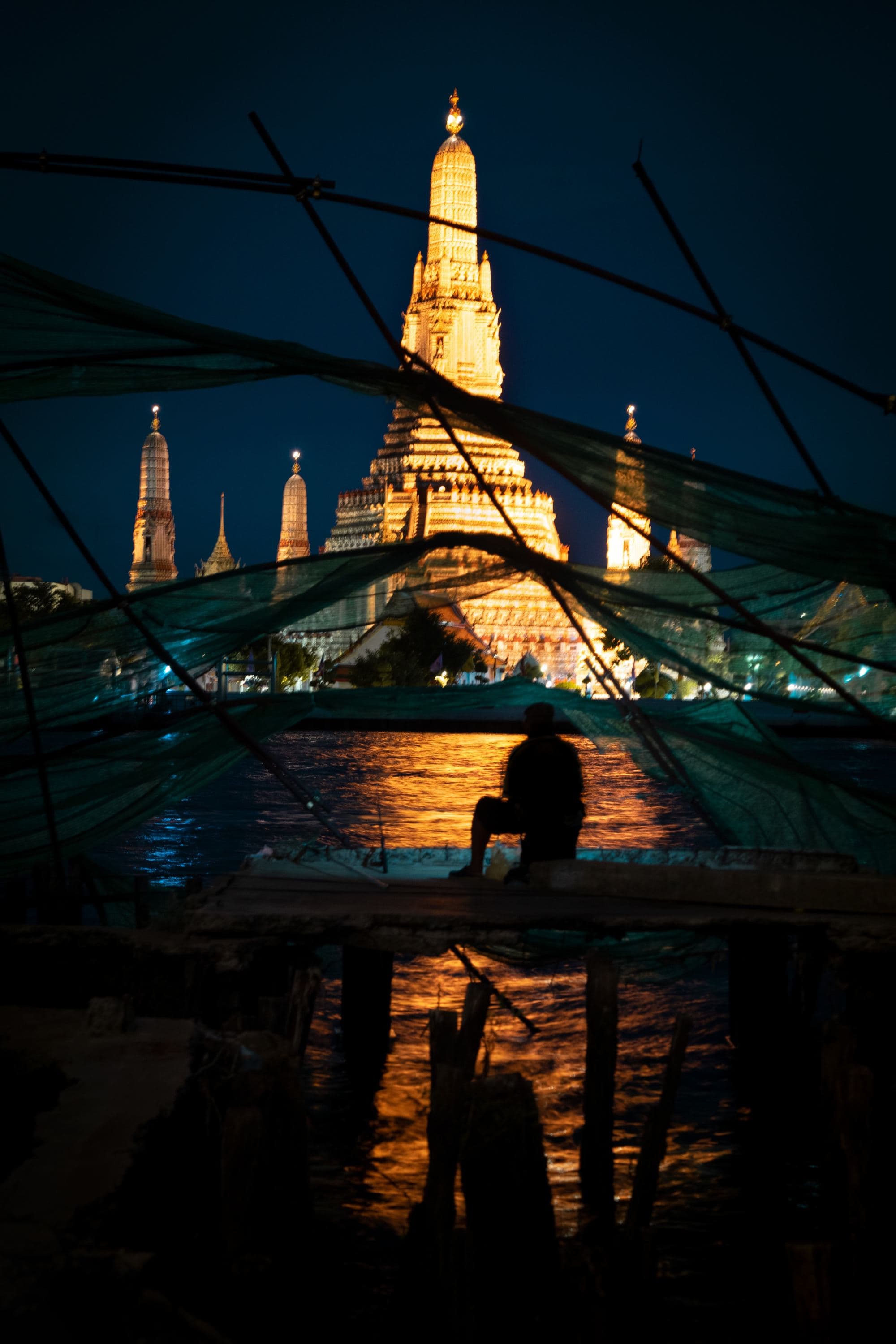 A person sitting on a dock at night in front of a light tower.