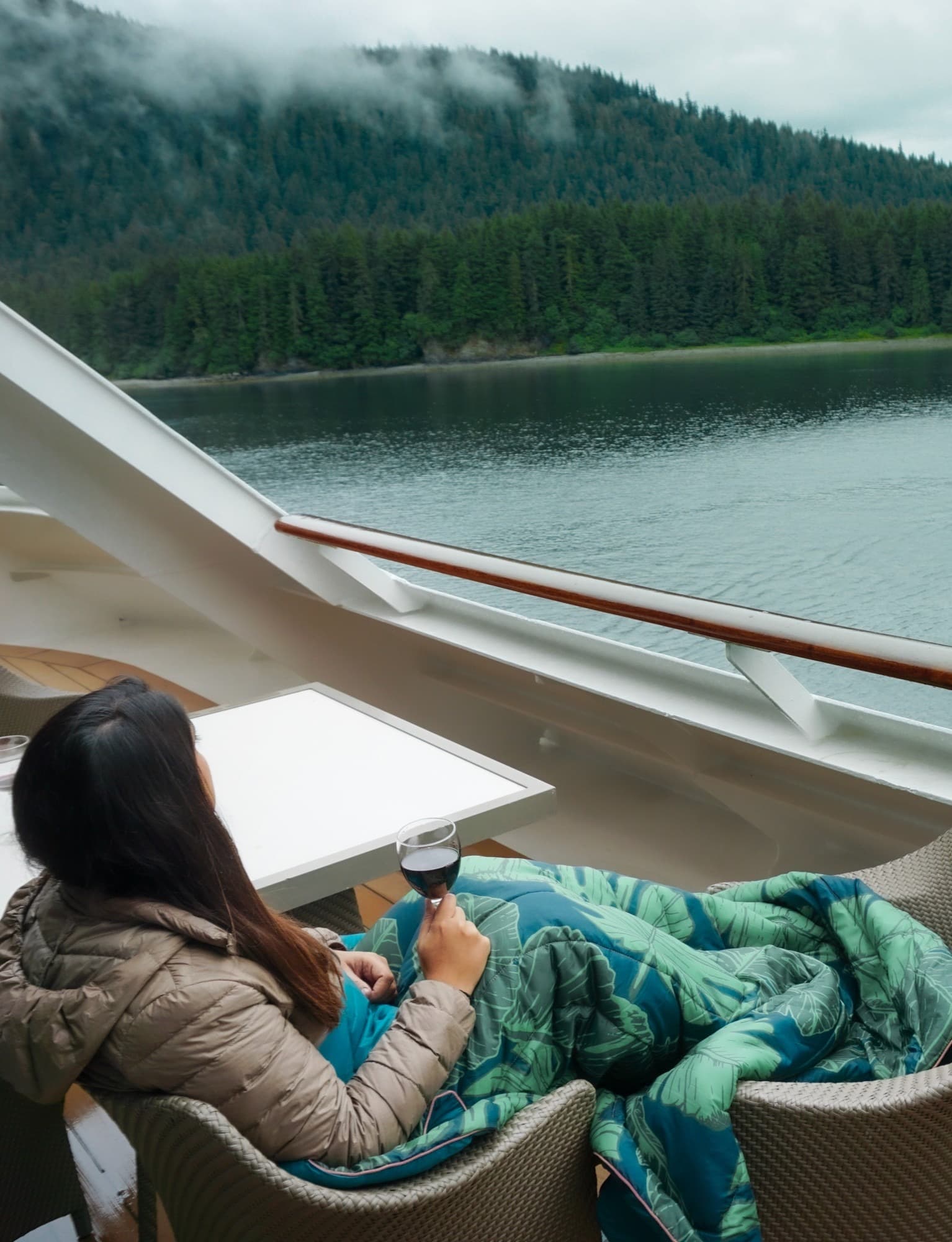 A person sitting on a deck chair on a cruise ship, wrapped in a blanket, looking out at a calm river and forested hills with misty clouds above.