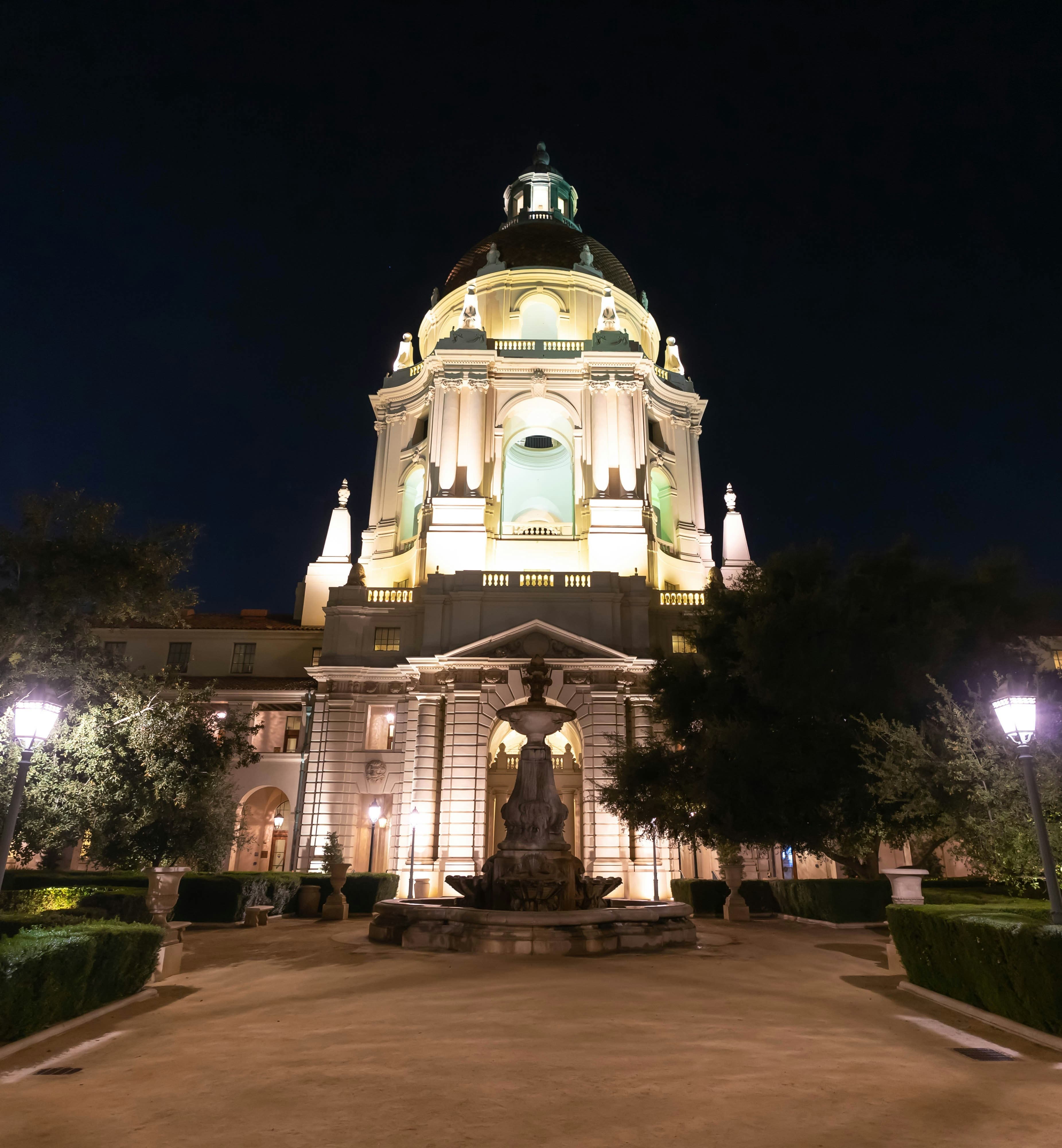 The picture depicts a large white building with a dome, a statue and trees lit up at night time. It is Pasadena City Hall.