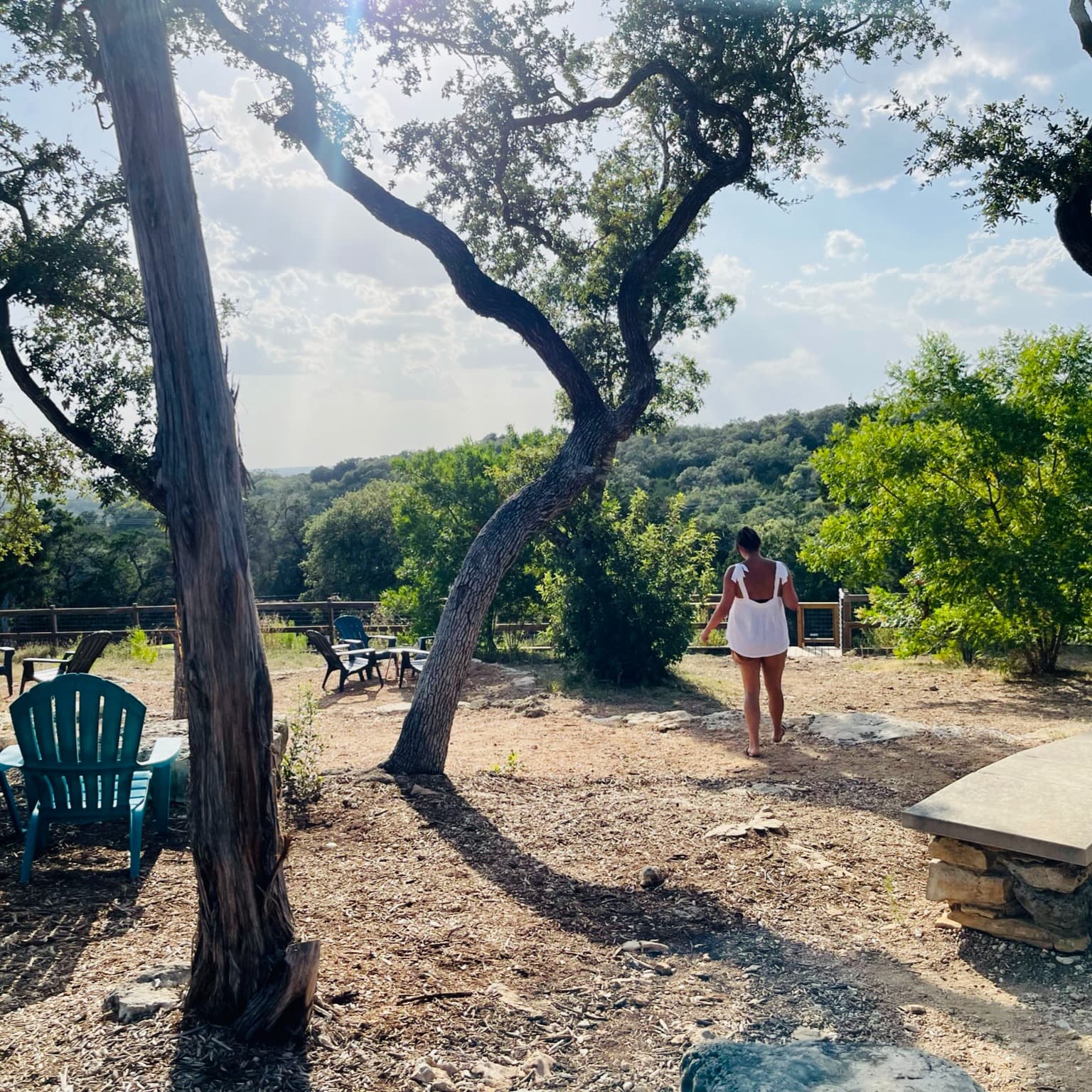 A woman in white dress in a park with trees and outdoor blue chairs.