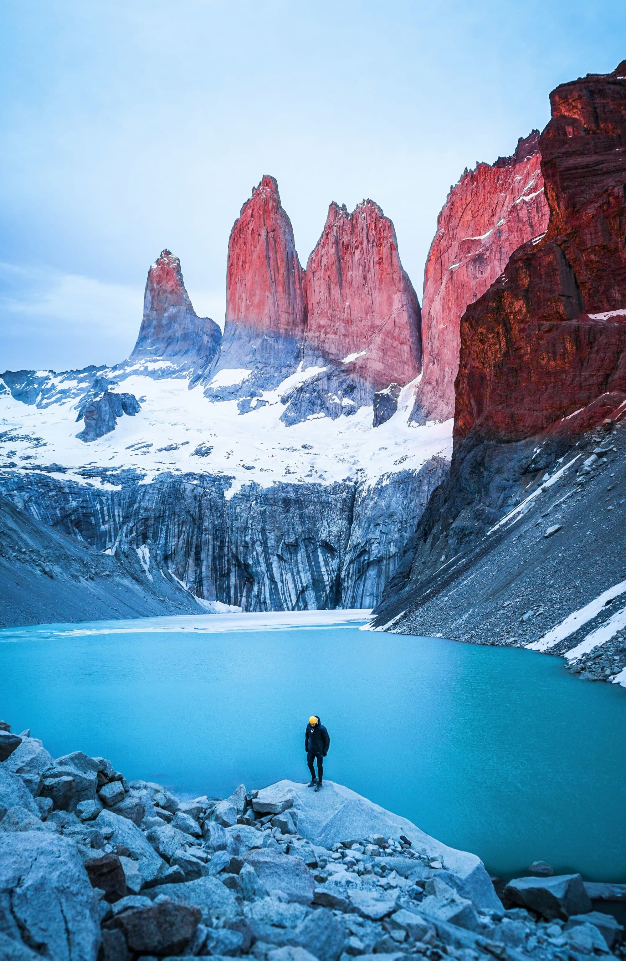 A traveler stands by a turquoise glacial lake at the foot of majestic red peaks partially covered in snow.