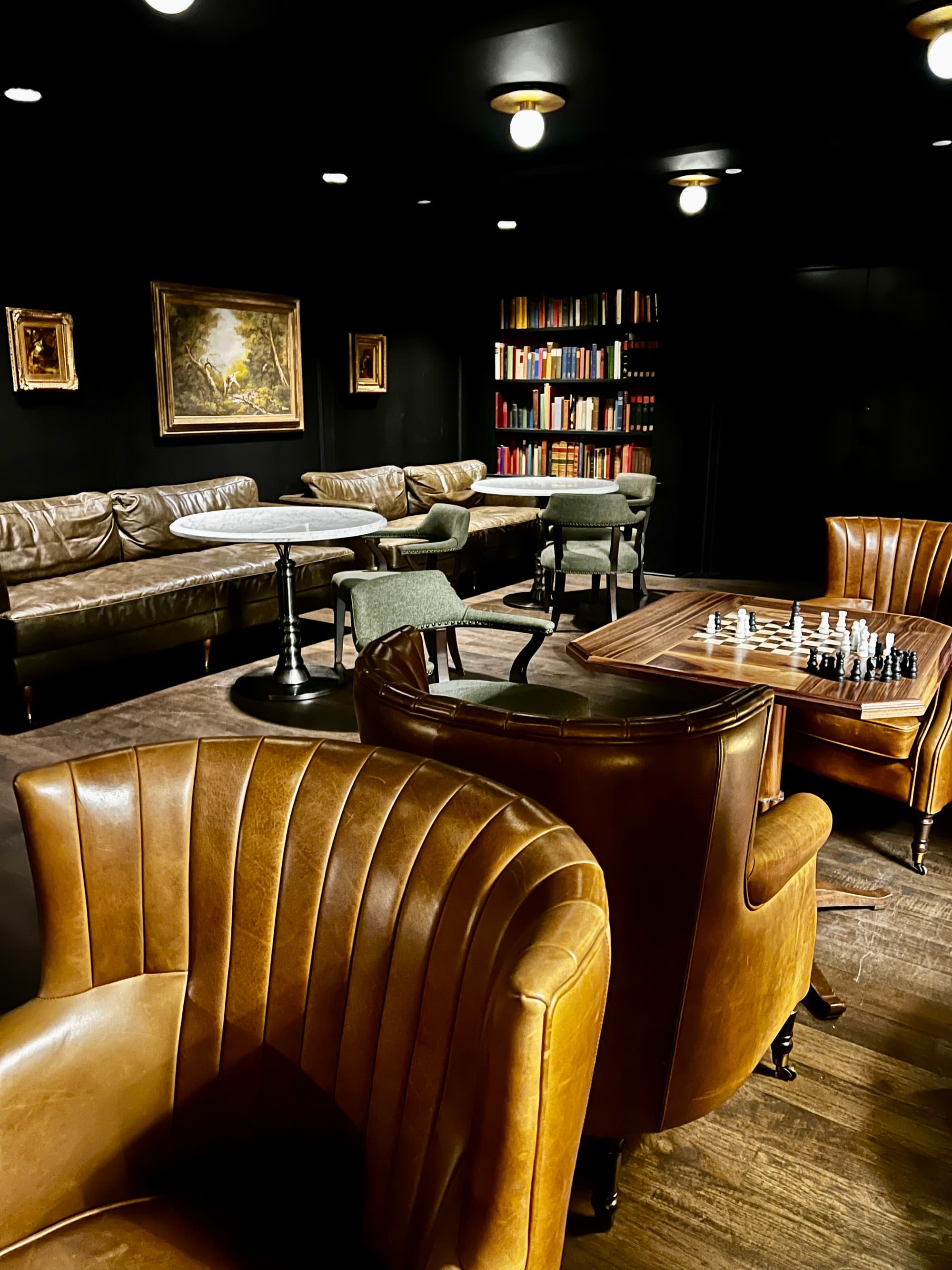 A view inside a dimly lit lounge with brown leather chairs, booths, tables, a chess board and bookshelves filled with books.