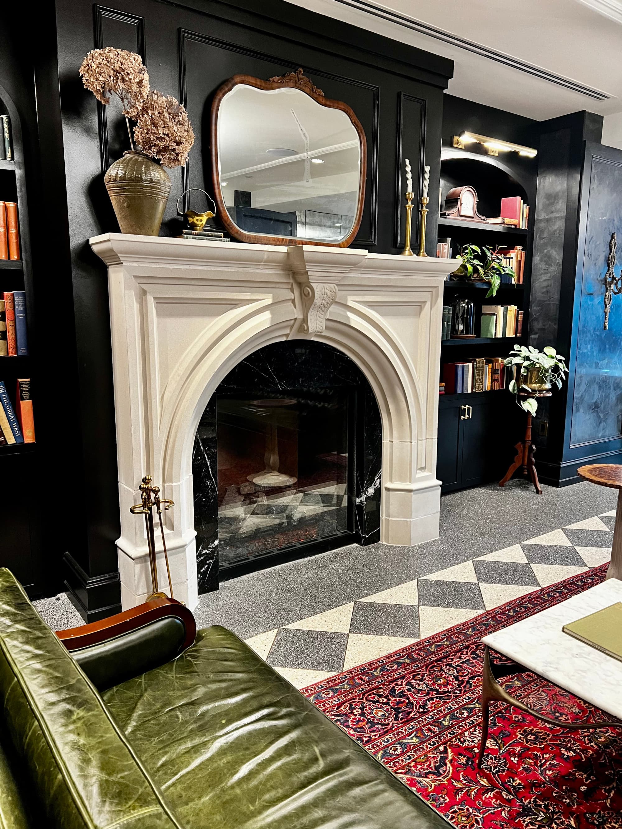 A room view of a white fire place, black walls, a large mirror on the mantel, diamond tiles in gray and white, a red rug with a table and a booth.