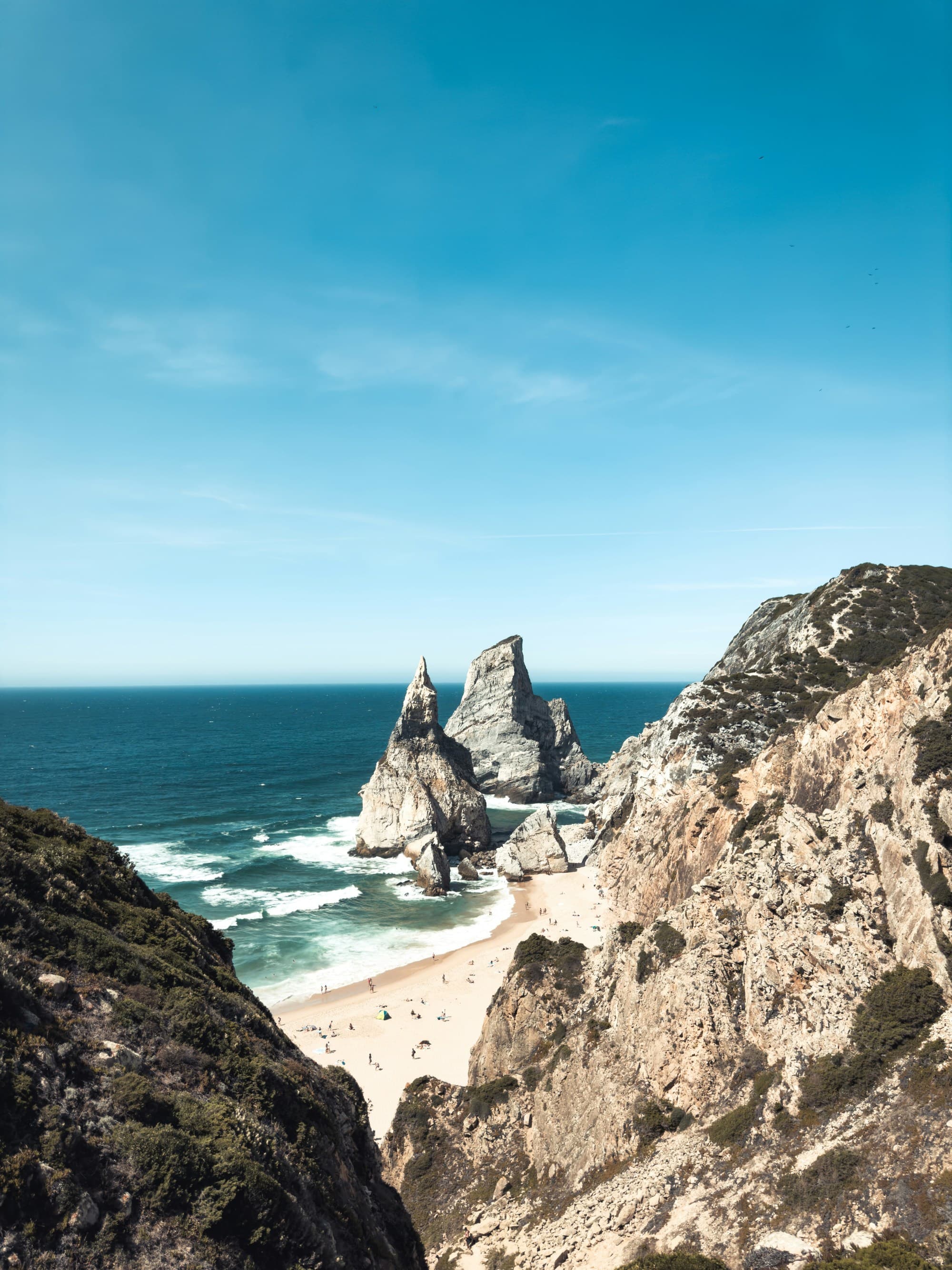 The image displays a coastal landscape with majestic rock formations on a sandy beach under a clear blue sky.