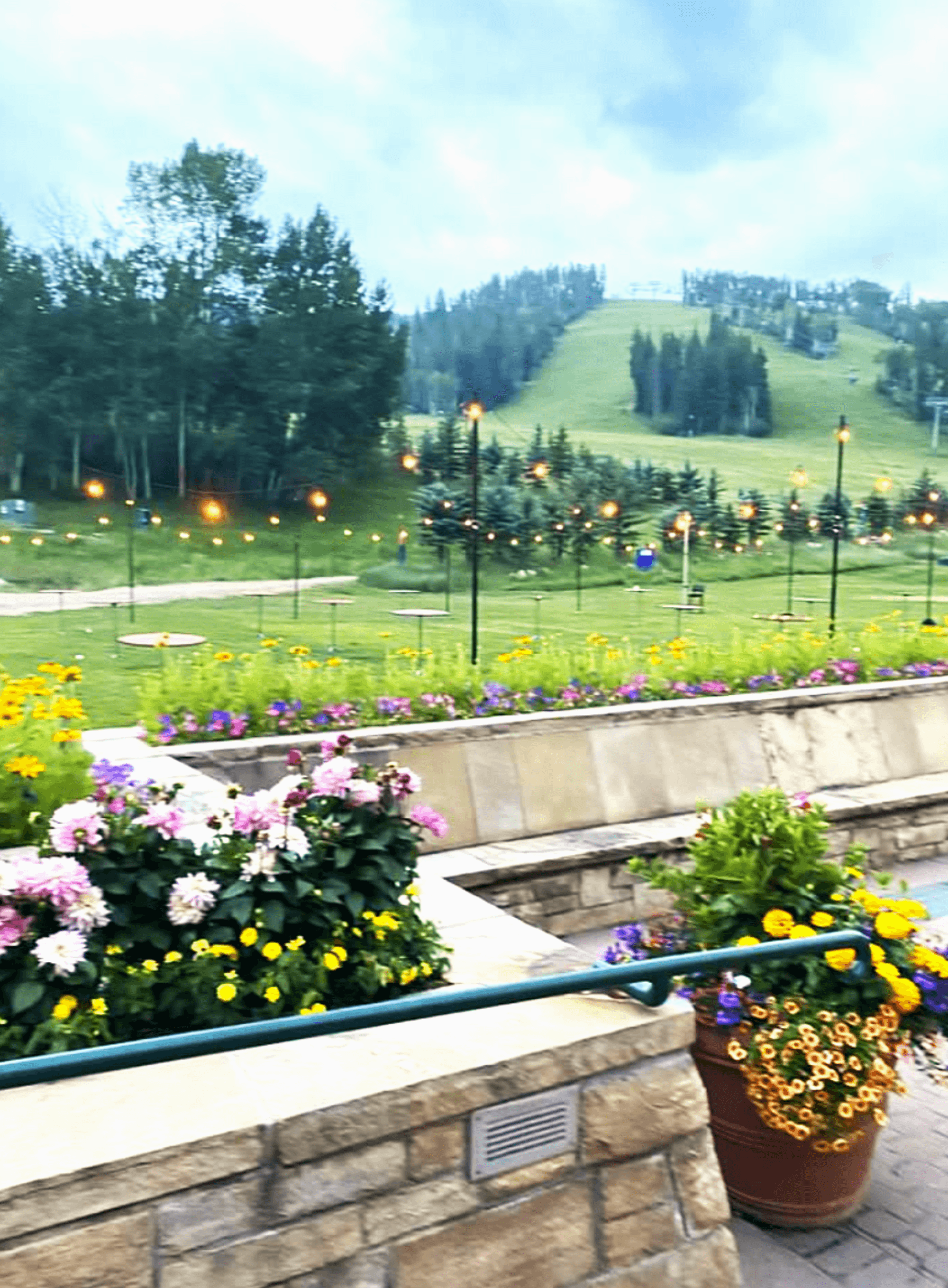 A picturesque park scene with colorful flowers, illuminated lamps, and a hilly backdrop under a cloudy sky.