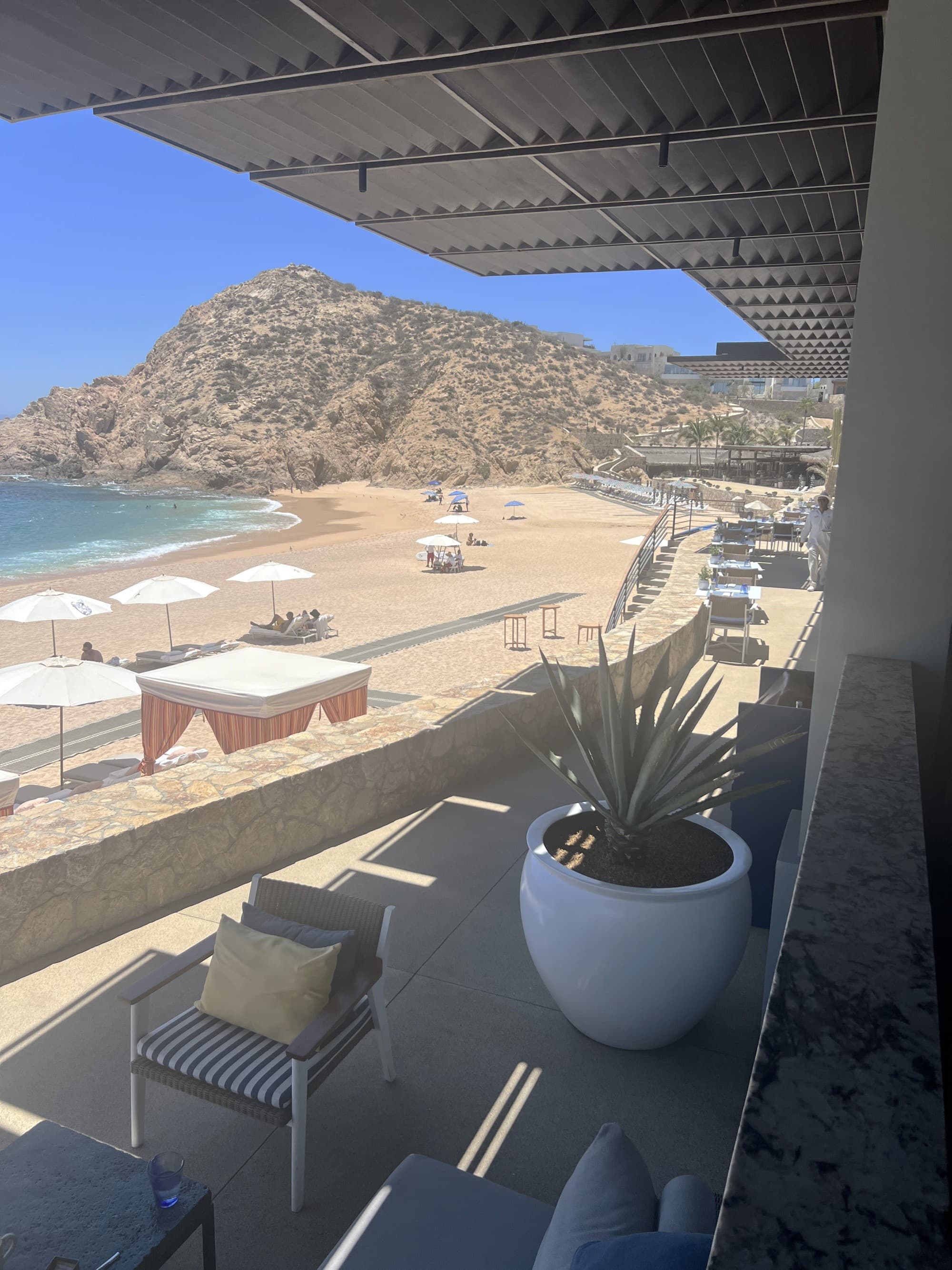 A tranquil beach view from a shaded balcony, complete with outdoor furniture and lush greenery.