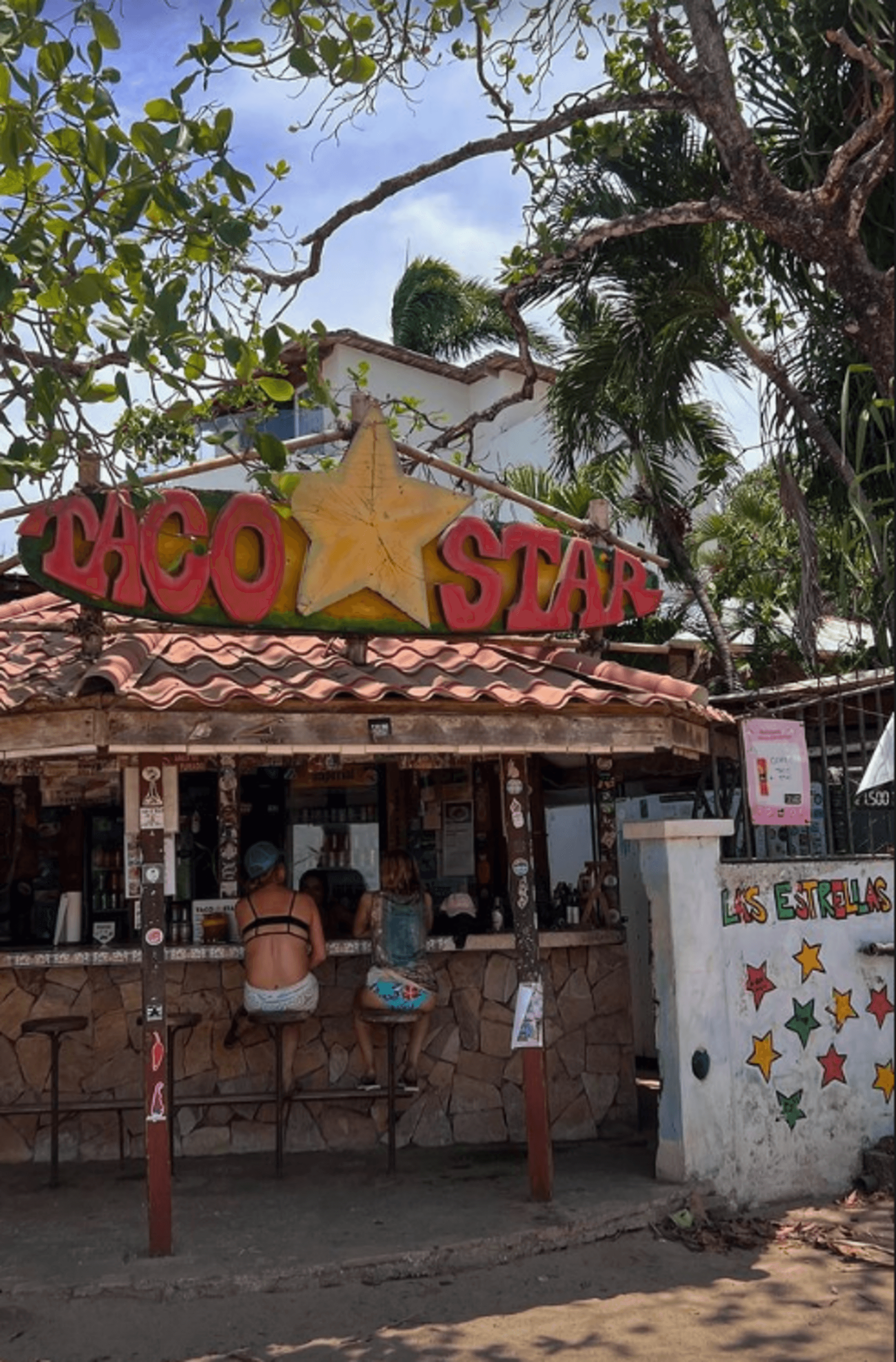 A bustling outdoor taco stand, “Taco Star,” serves patrons under a colorful sign and tropical foliage.