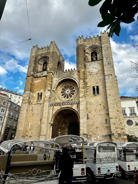 An exterior view of Lisbon Cathedral, built with Romanesque and Gothic architecture. There are small electric carts parked in front of the building.