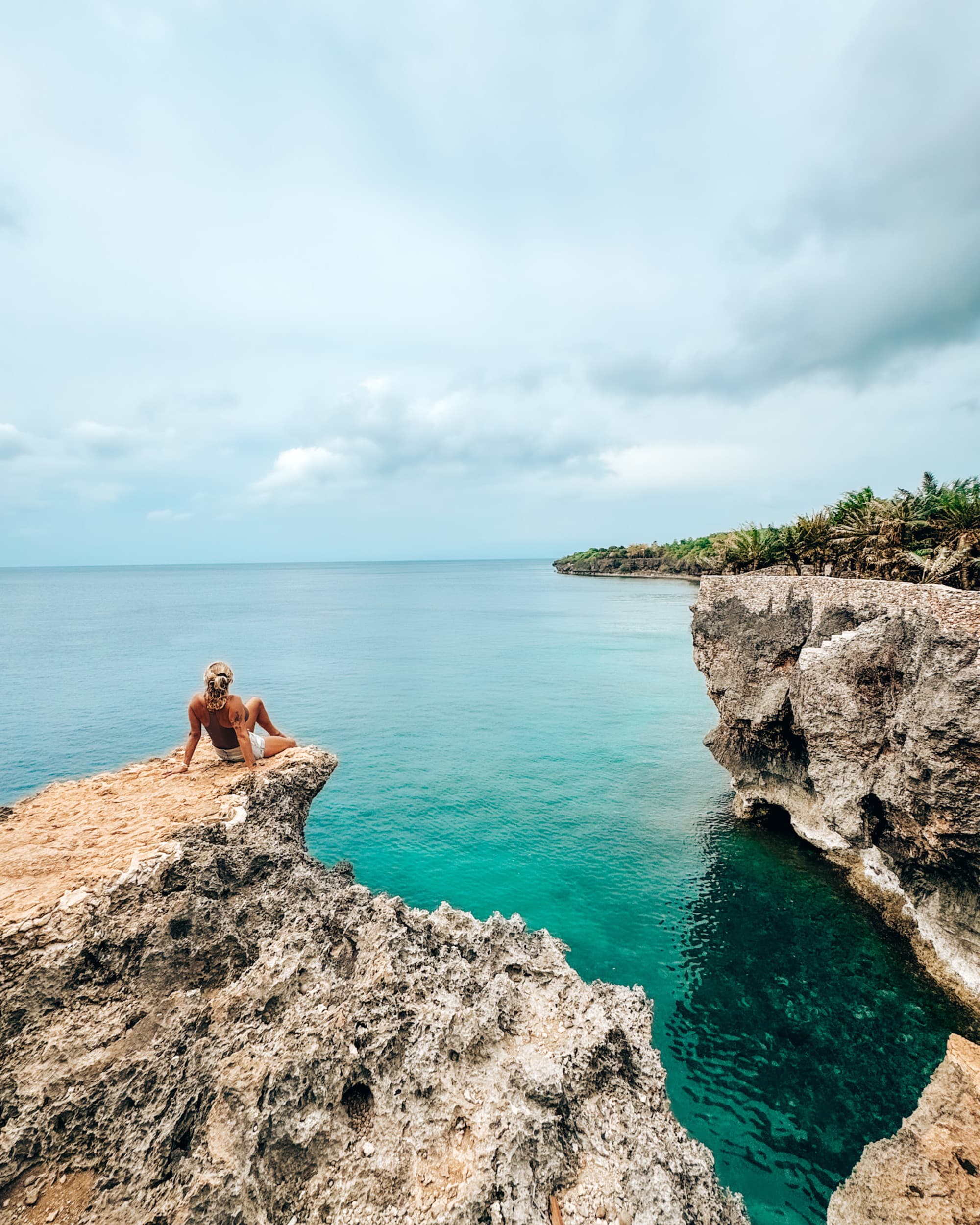 A picture of a person sitting on a rocky cliff near the water during the daytime.