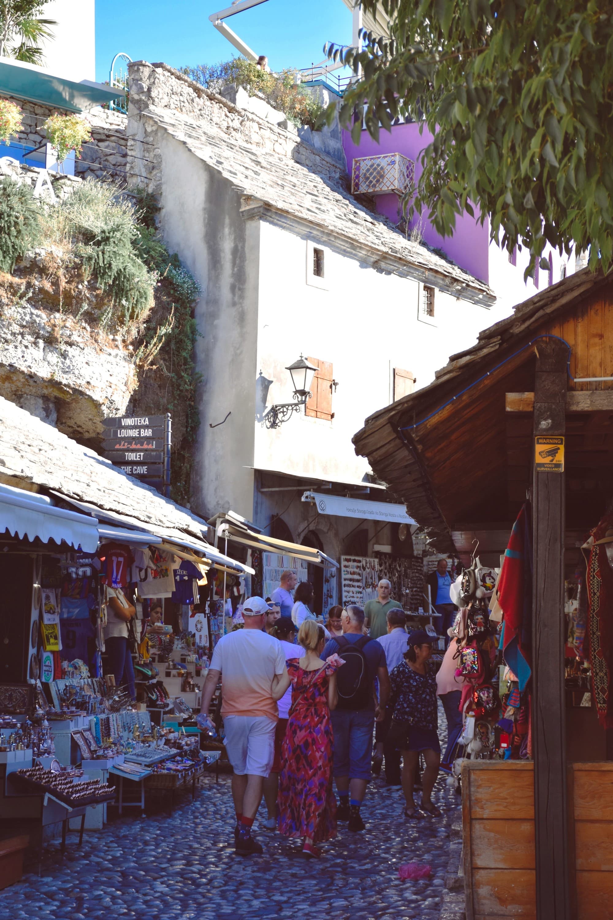 A picture of people walking down a cobblestone street with various markets, stands and shops surrounding them on the left and right sides. There is a white building with a slanted roof in the distance, and trees near it.