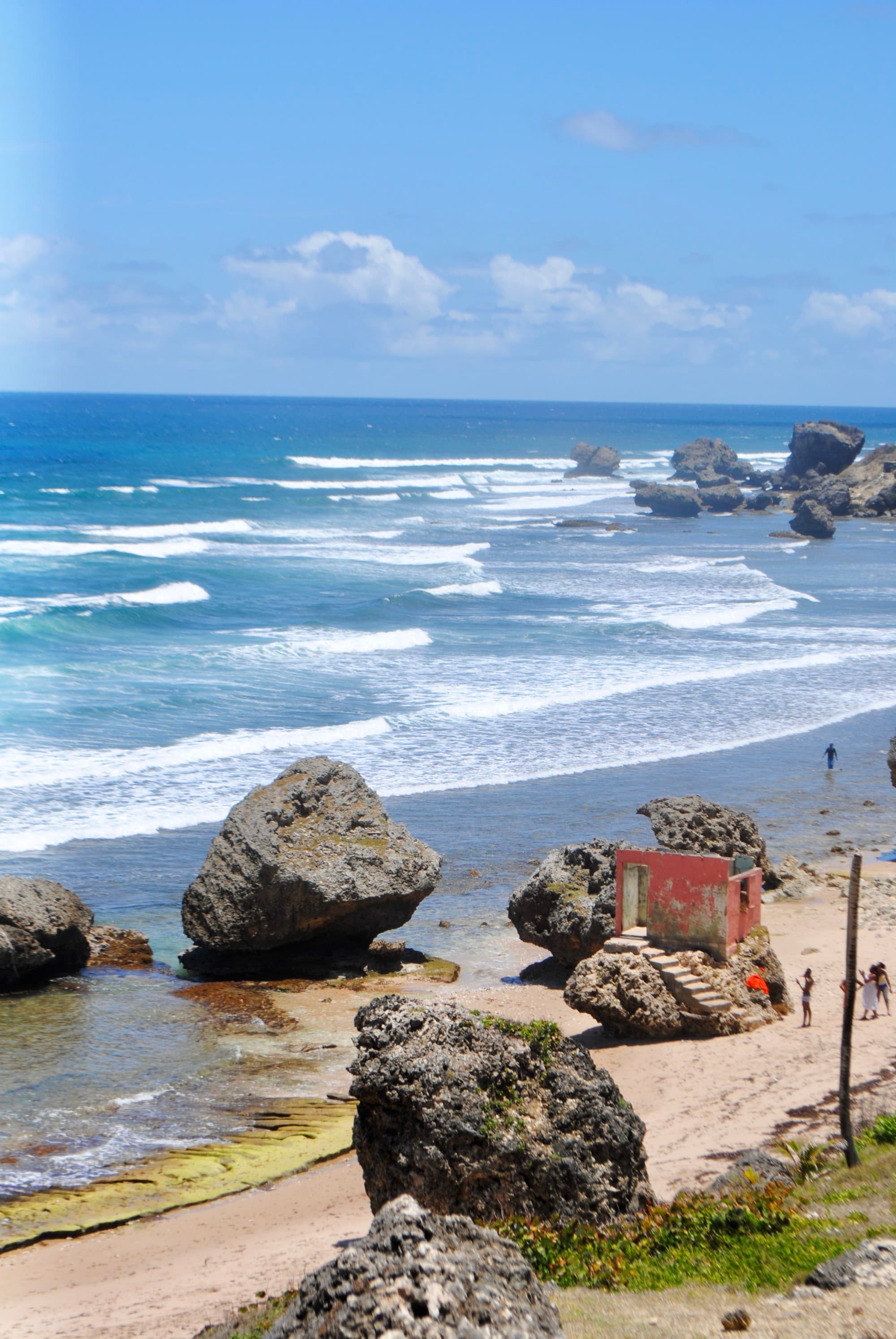 A view of the shore littered with big stone boulders, bright blue water and a blue sky.