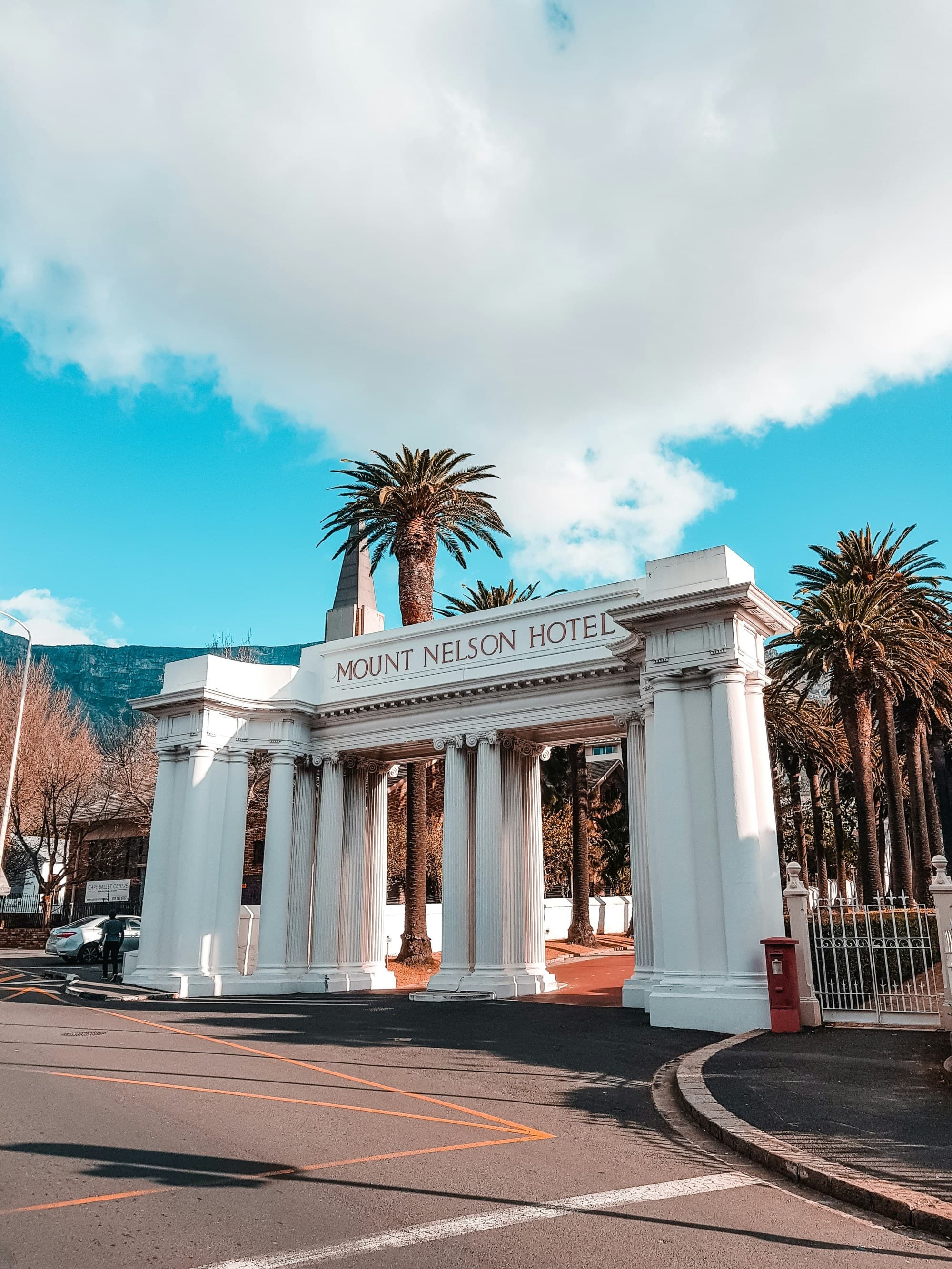 A beautiful entrance with white arches to the Mount Nelson Hotel, surrounded by palm trees and a road.