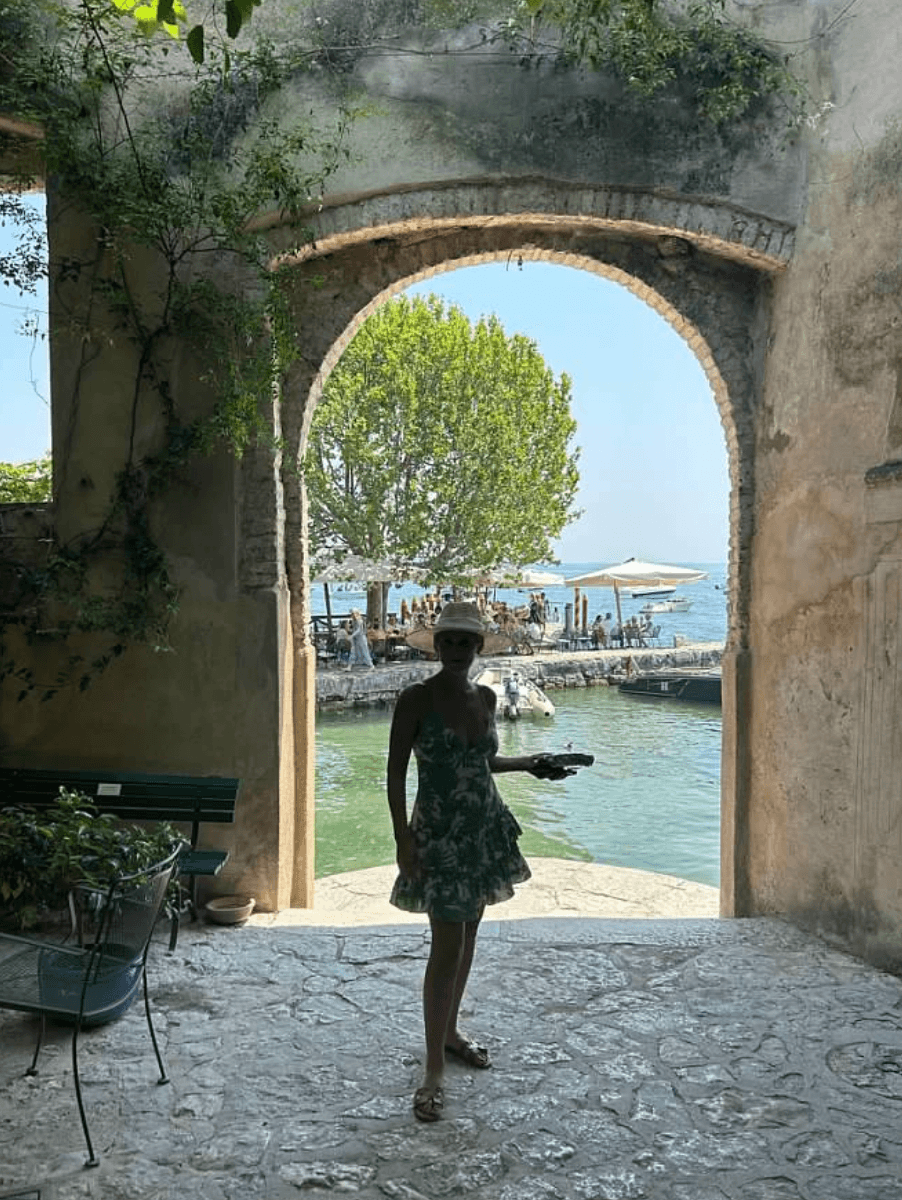 A woman posing for an image in front of a stone archway with a view of a tree, water and patio full of people dining under umbrellas.