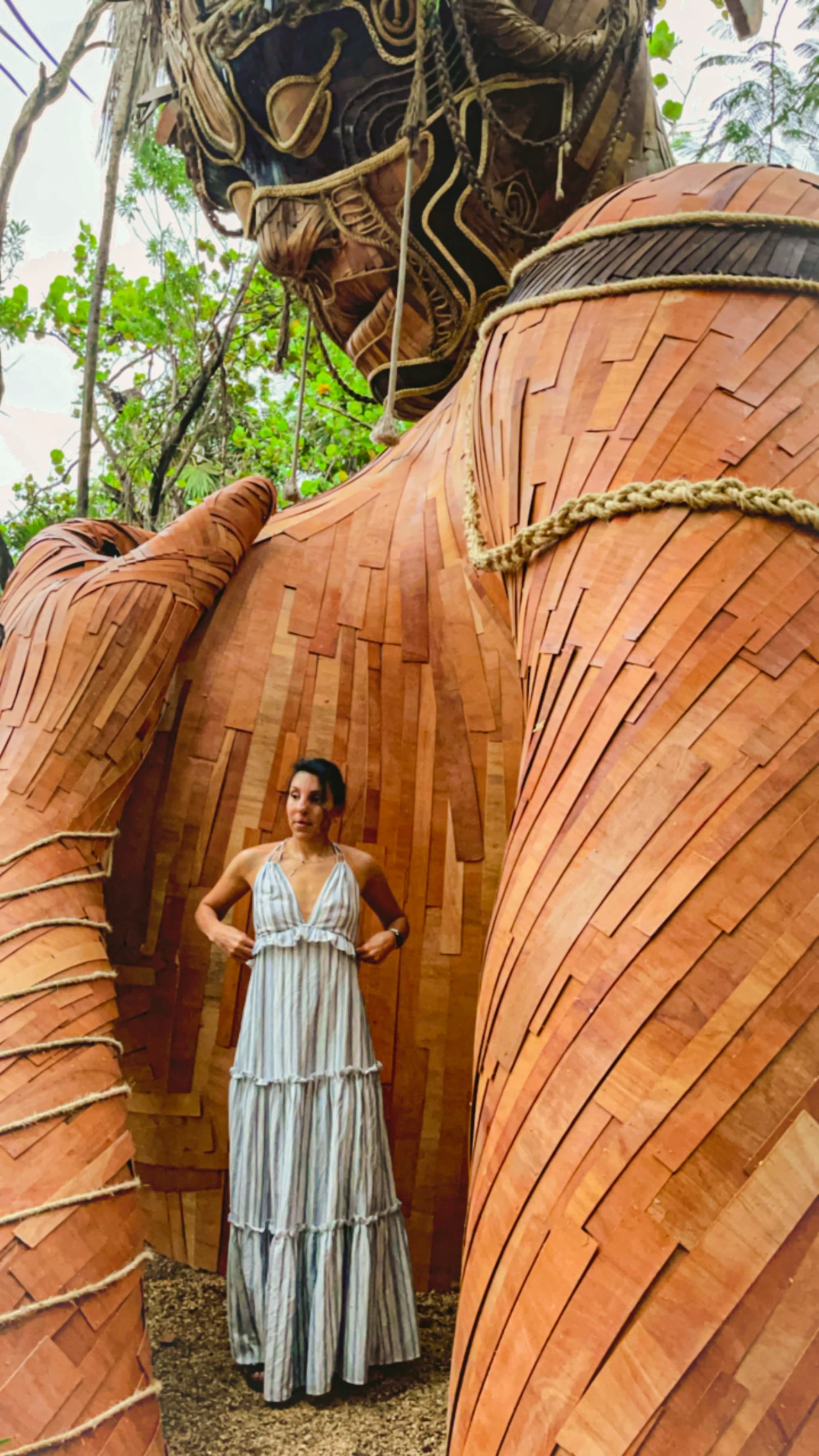 A woman posing near a huge wooden sculpture during daytime.