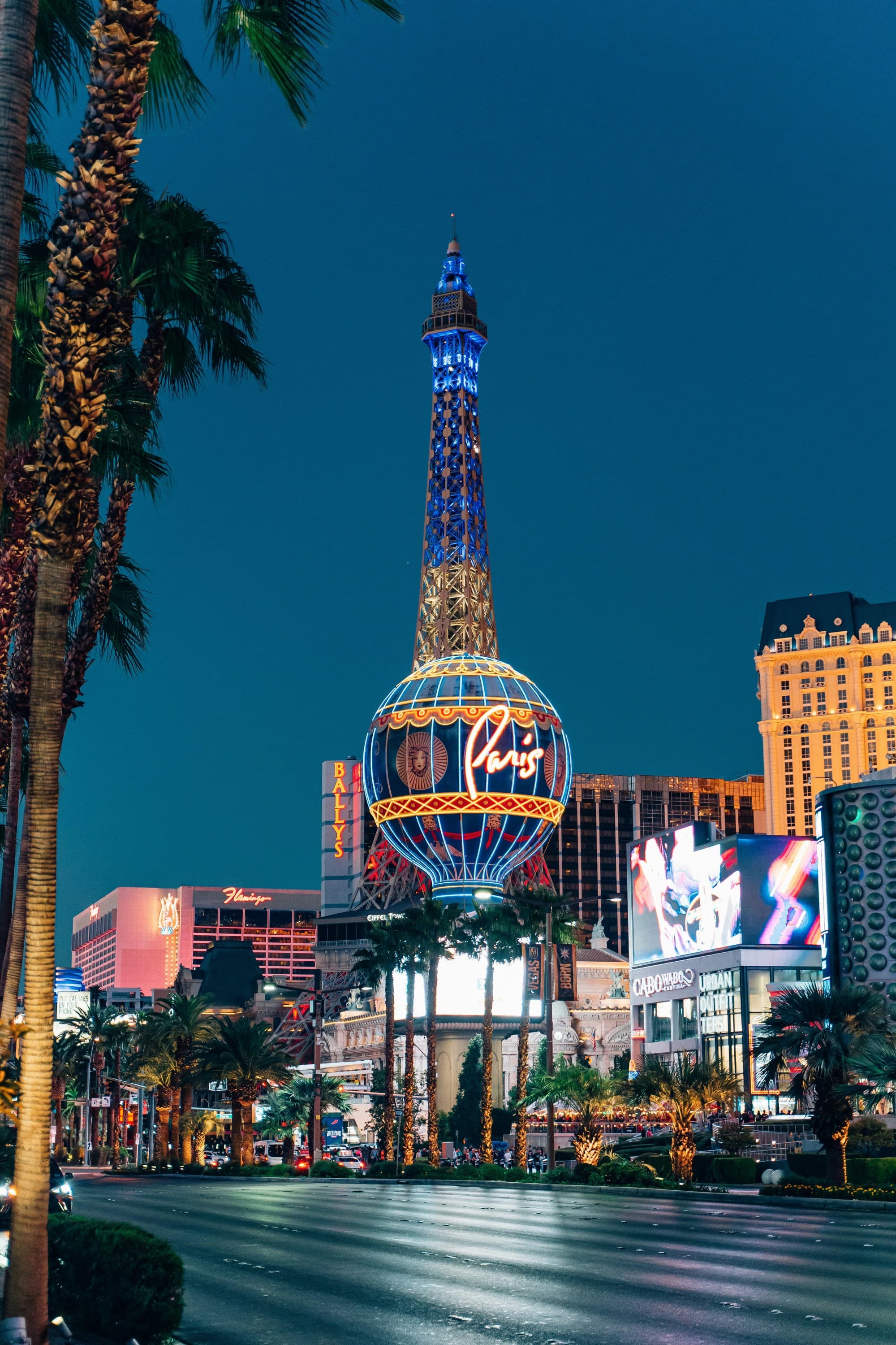 A picture of the Eiffel Tower in Las Vegas at night surrounded by buildings, cars and palm trees.