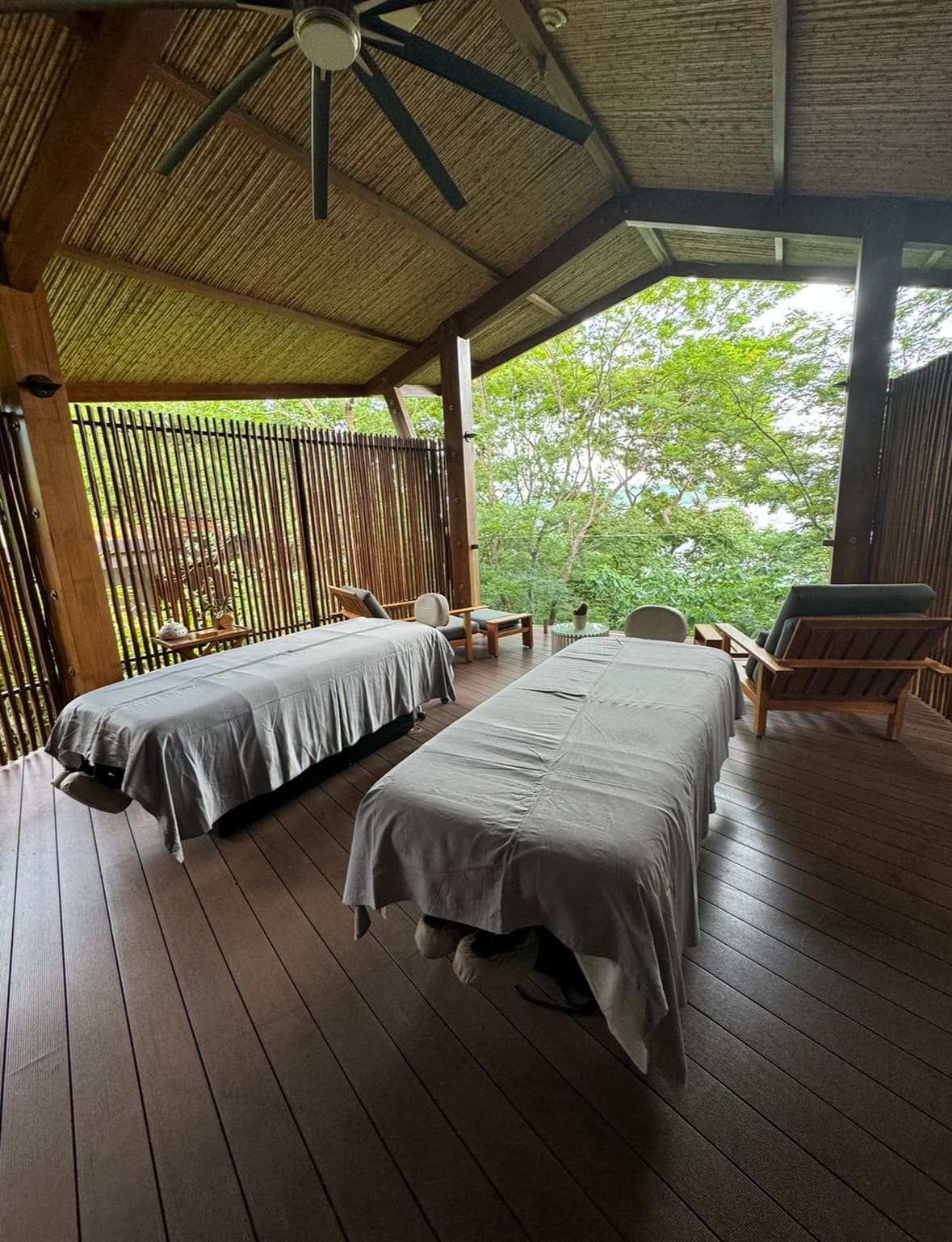 A view of the Andaz Spa couples massage area with two tables set under a teak roof surrounded by lush greenery.