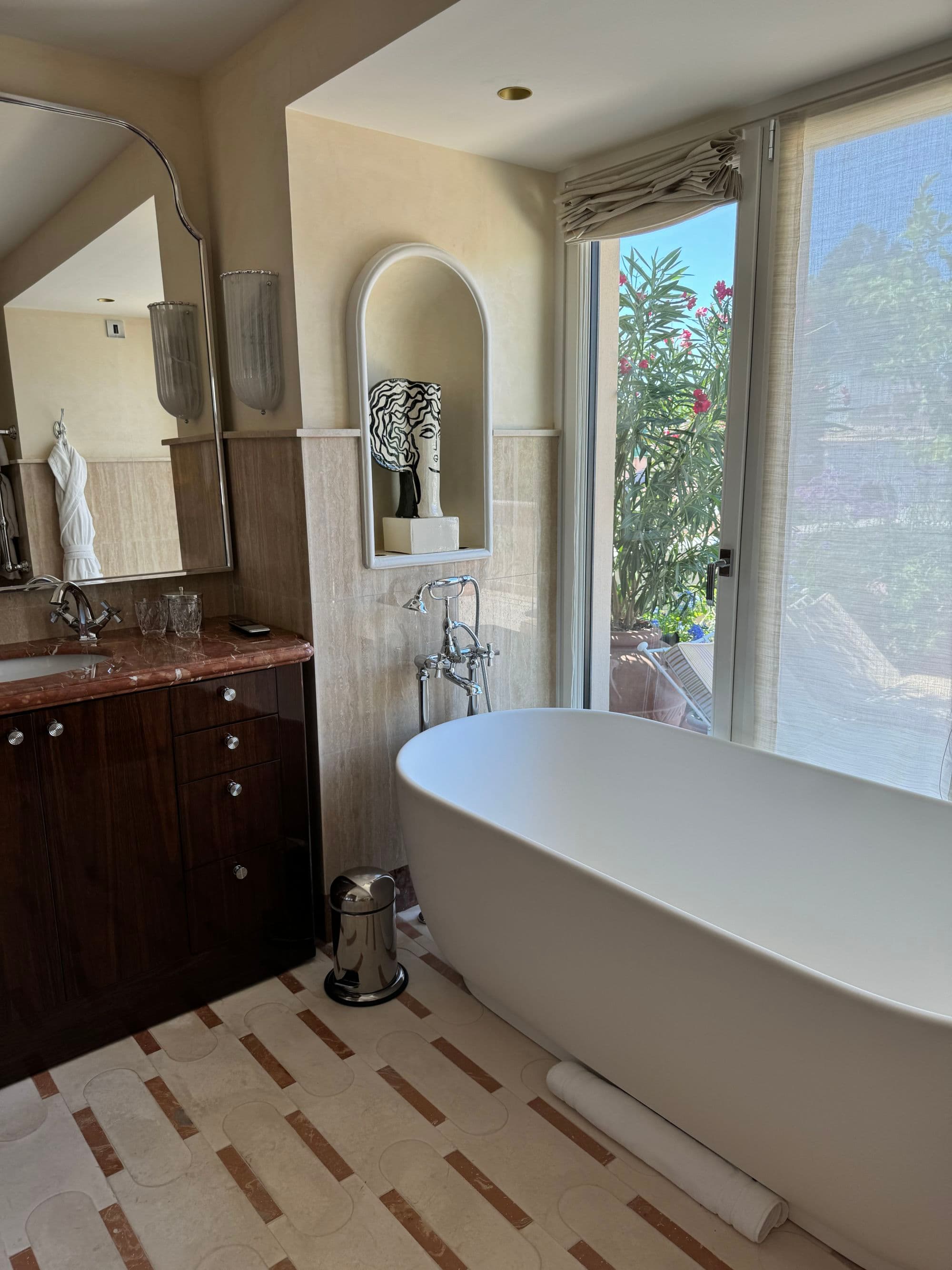 The hotel bathroom is spacious with tiled floors and a large tub and vanity next to a window overlooking a garden terrace.