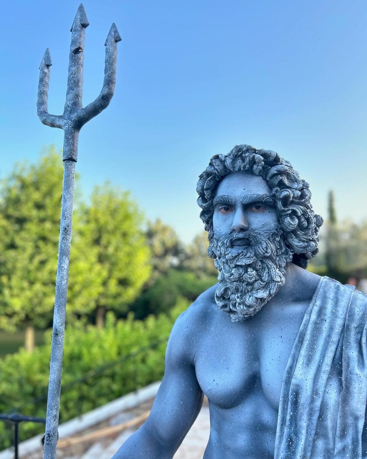 A person painted to look like a Greek statue holding a trident standing in front of greenery at the welcome dinner - Kristyne Wada