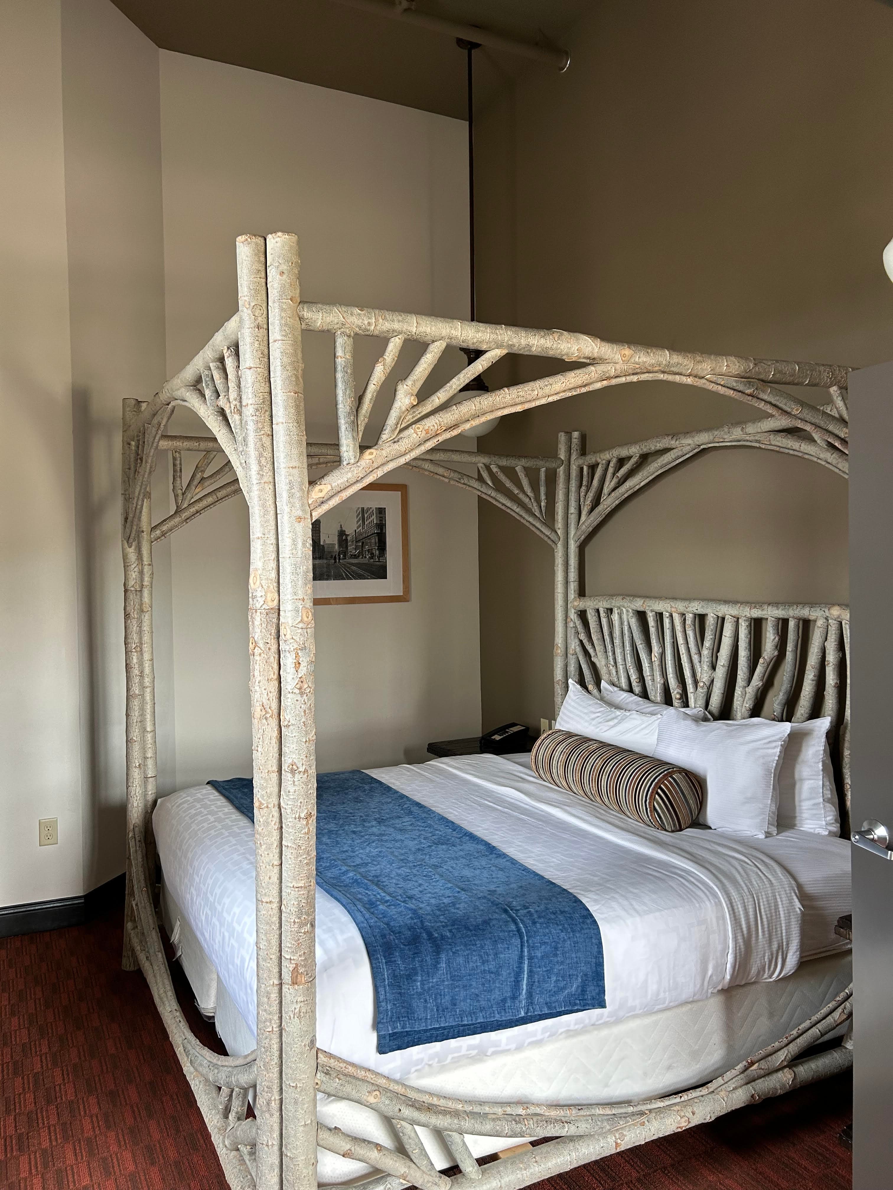 Hotel room bed made up under a wooden four-poster canopy made of branches.