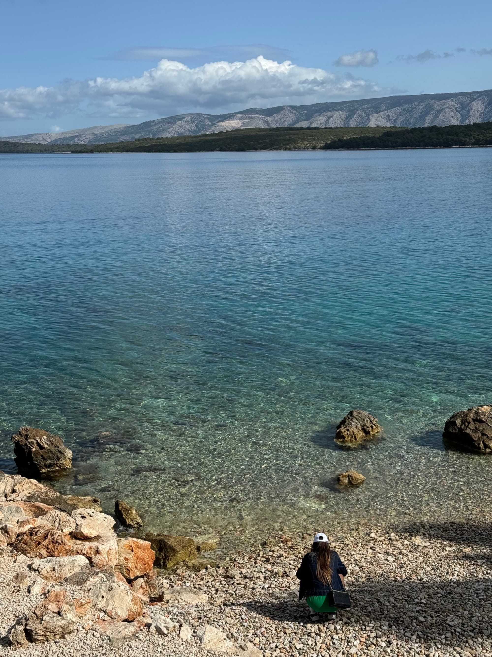View of crystal clear coastal waters with rolling hills unfurling in the distance on a clear day dotted with clouds.