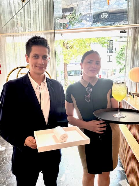 Two people posing for a photo with a towel on a tray and a yellow iced beverage in a wine glass on another tray. They're wearing fancy clothing, and there is a window with a tree and other buildings in the background.