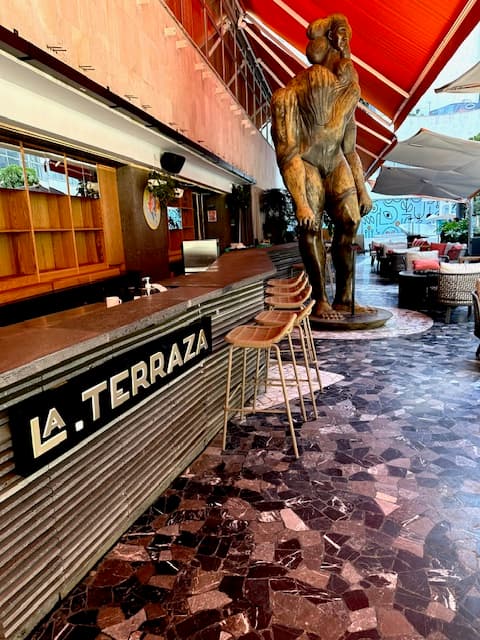 This image depicts a view of a large human-like statue made out of wood, tiled flooring, bar stools, a counter top and wood shelving. There is seating in the far right corner of the image, and a sign that reads "La Terraza" in white lettering on the bottom left side of the bar.