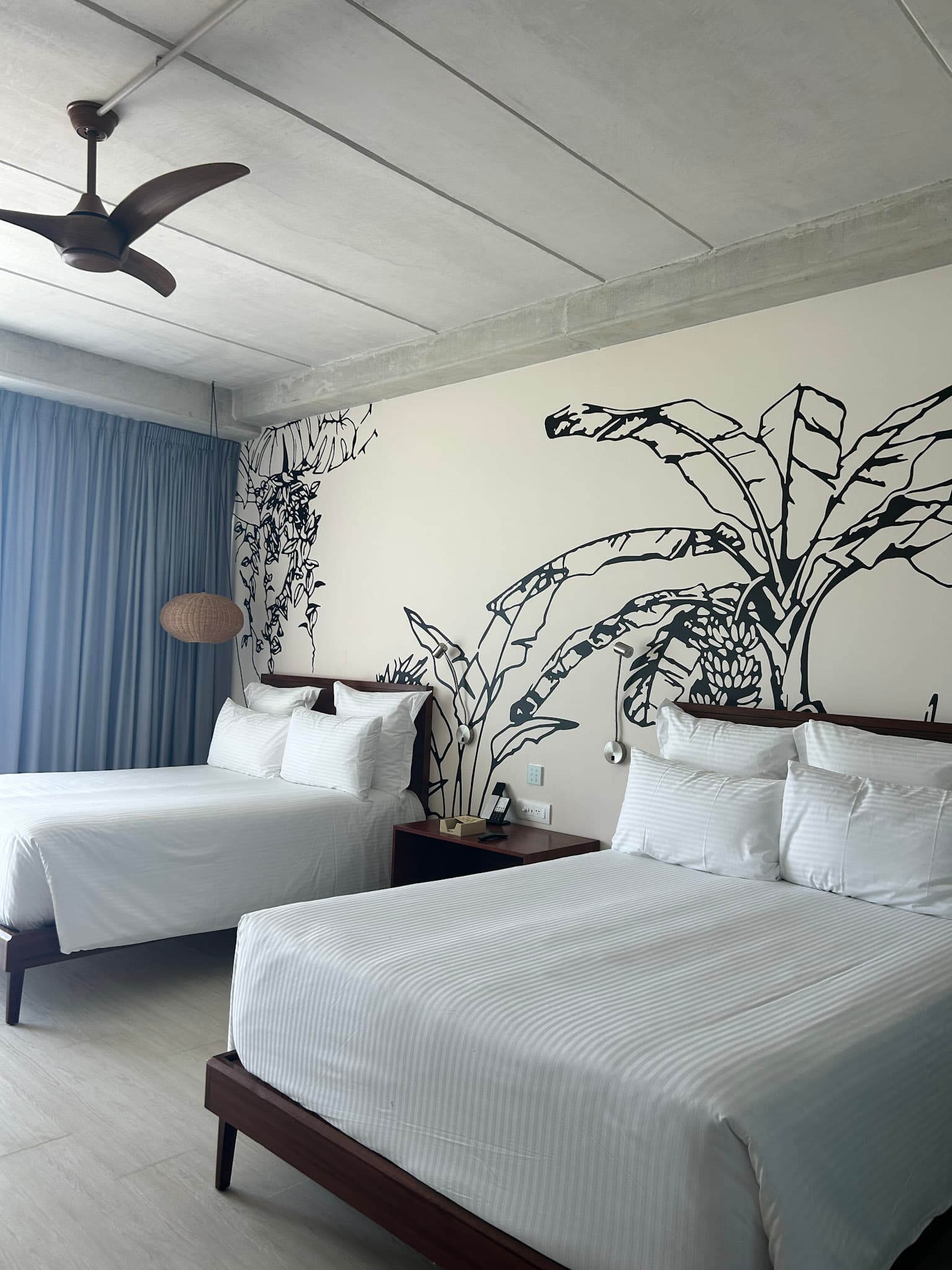 Inside a double queen bed room, with crisp, white bedsheets, a ceiling fan, and mural of palm trees on the wall.
