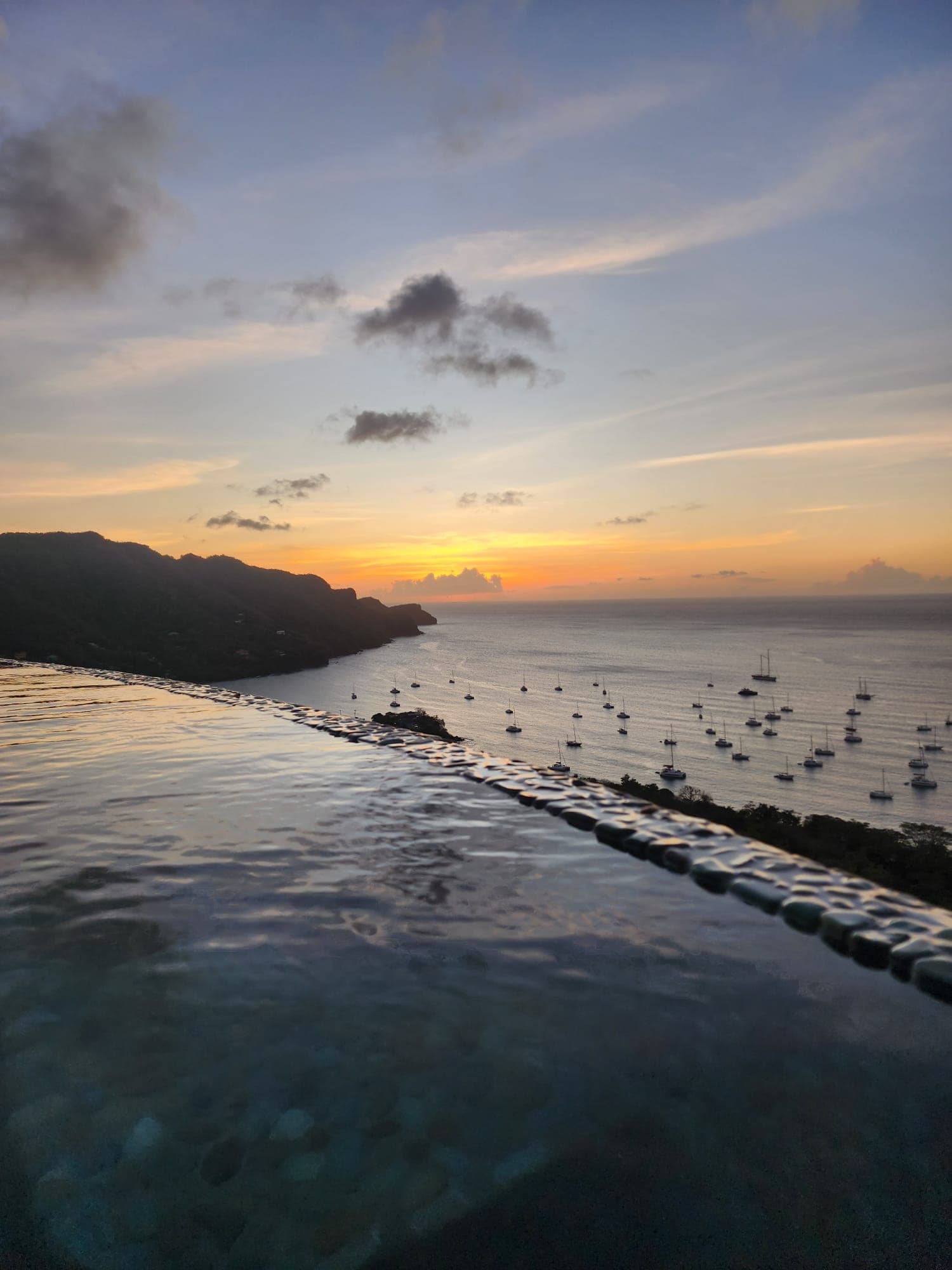 The orange and yellow sunset over the harbor and infinity pool. There is land and various boats in the surrounding area.