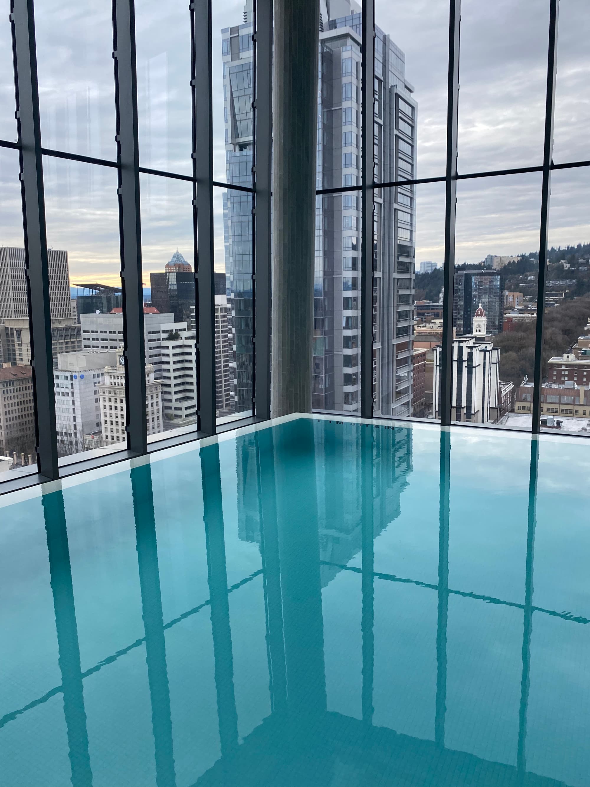A swimming pool with floor to ceiling windows looking out to a view of the city during the daytime.