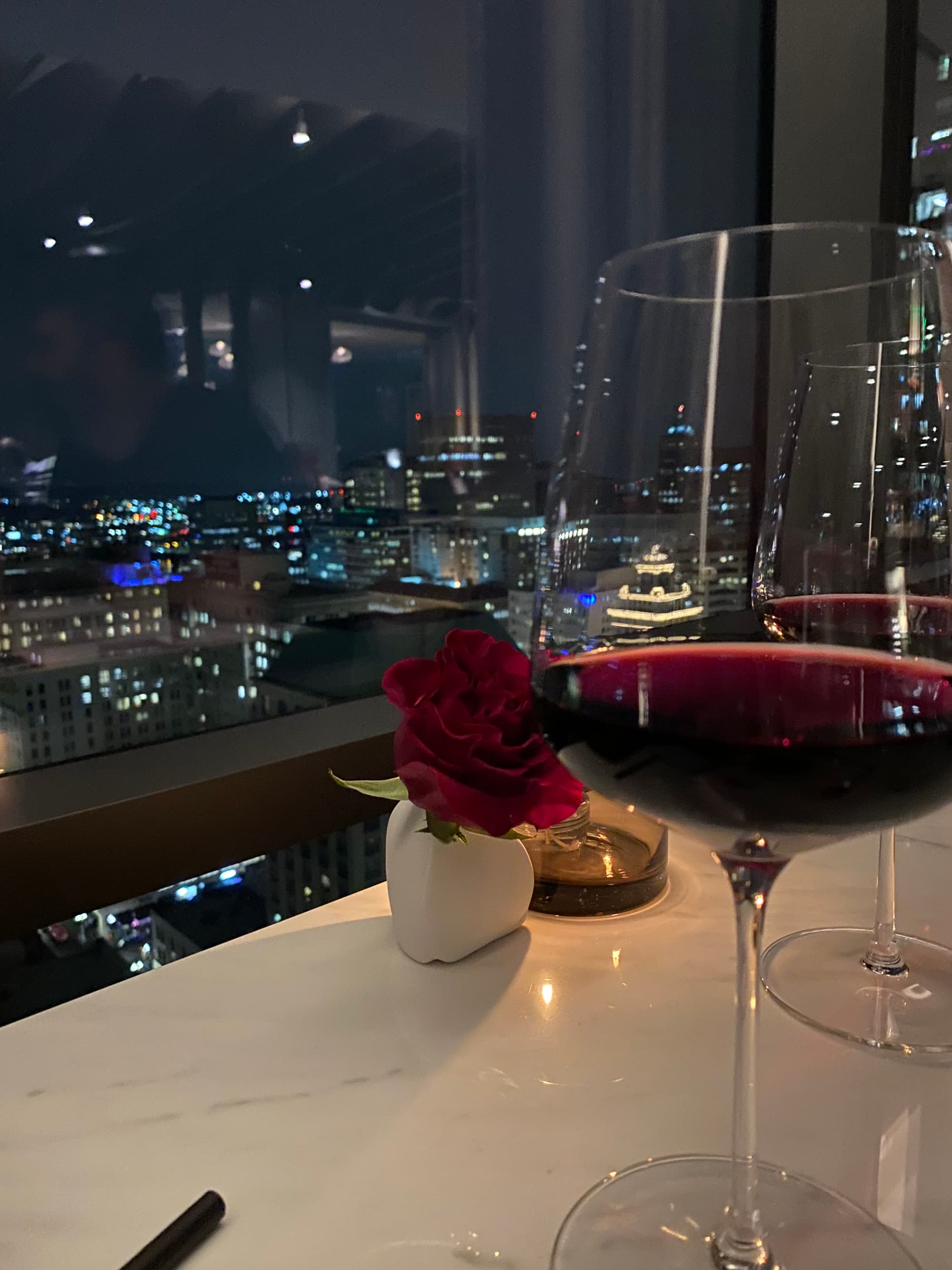 A dining table with a glass of red wine, white vase with a red rose and a view of the city skyline at night.