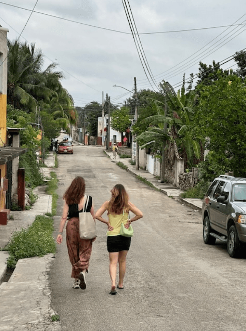 Two women walking down a road surrounded by fencing, palm trees and a parked car on the right.