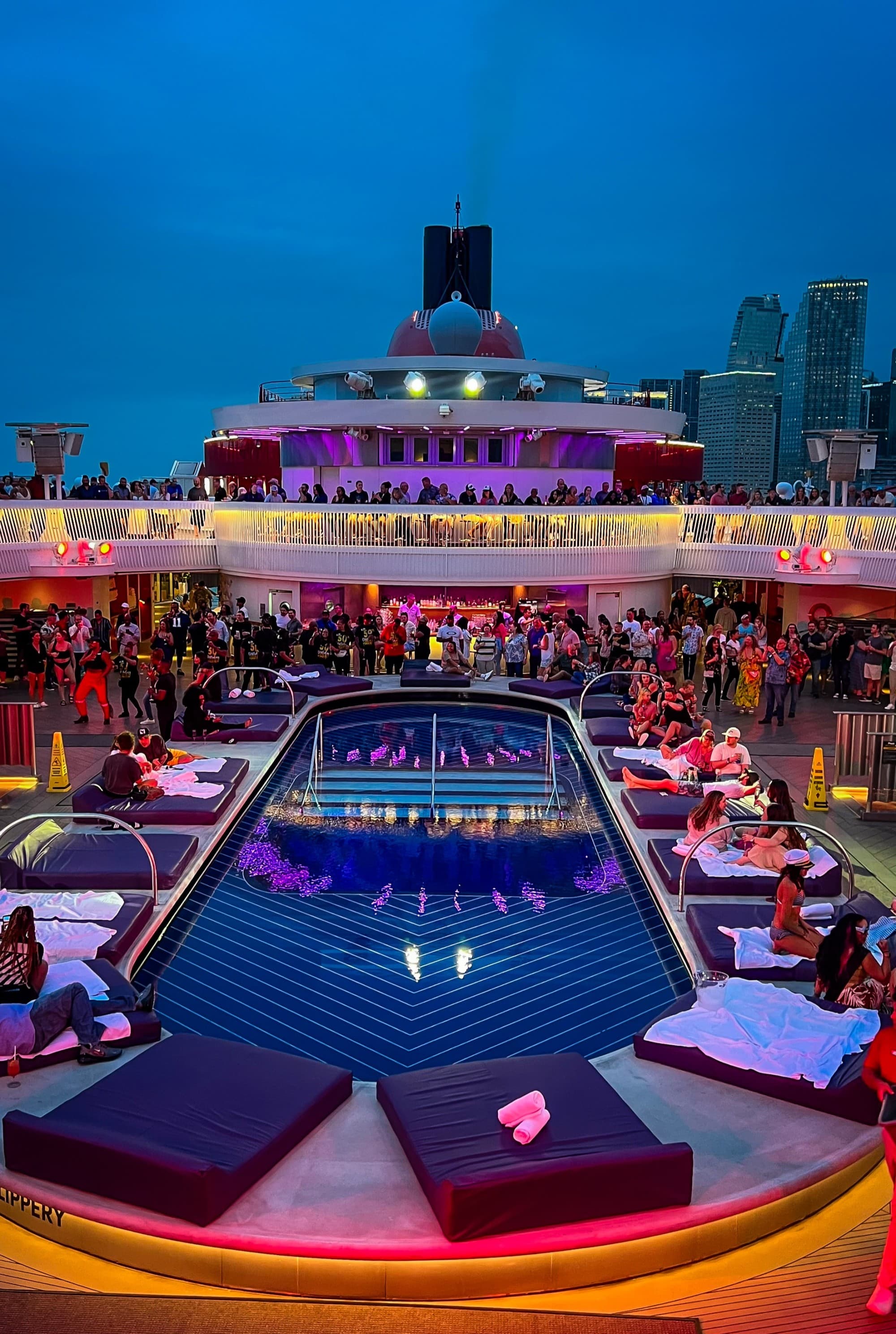 A pool on a lit-up cruise deck with lounge pads surrounding in the evening