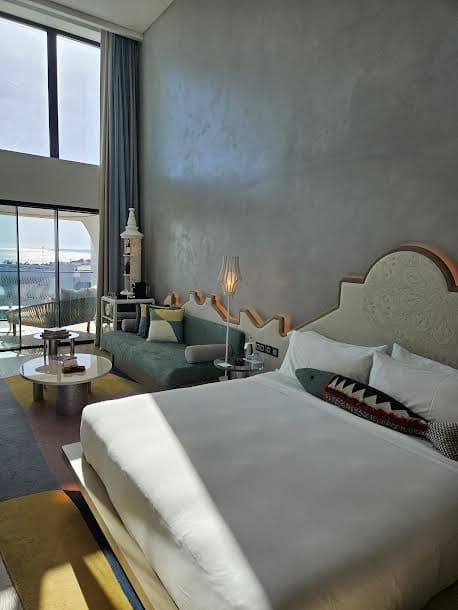 A view of the WOW Suite complete with luxurious white bedding, grey plastered walls and a couch with a decorative coffee table next to floor to ceiling windows and paneled curtains.