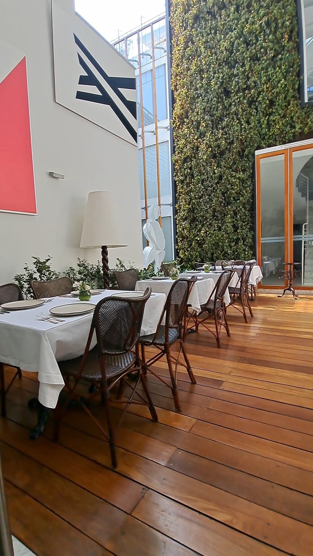 A dining area with white tables, black chairs, artwork, a foliage wall and a shiny wooden floor.