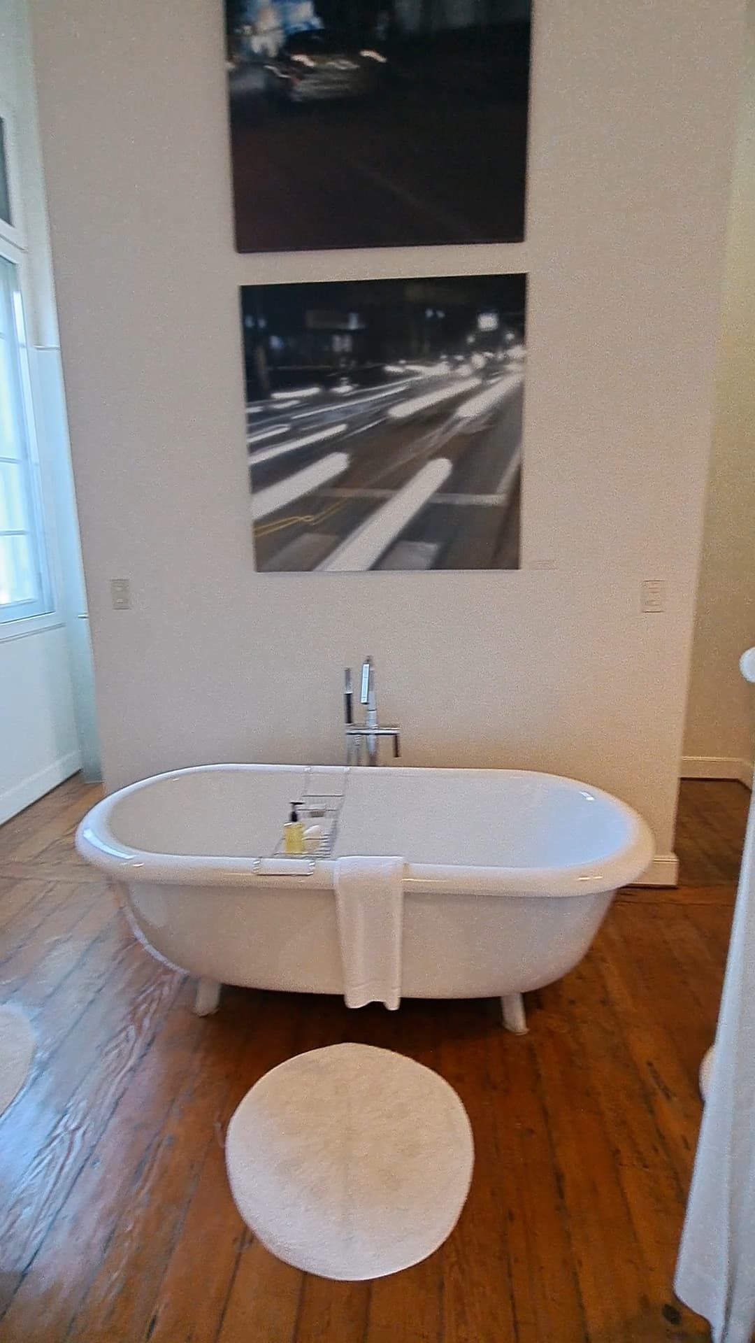 A white bathtub, white rug, towel and two pieces of artwork hanging on the wall in a bathroom. The floor is wood paneled.