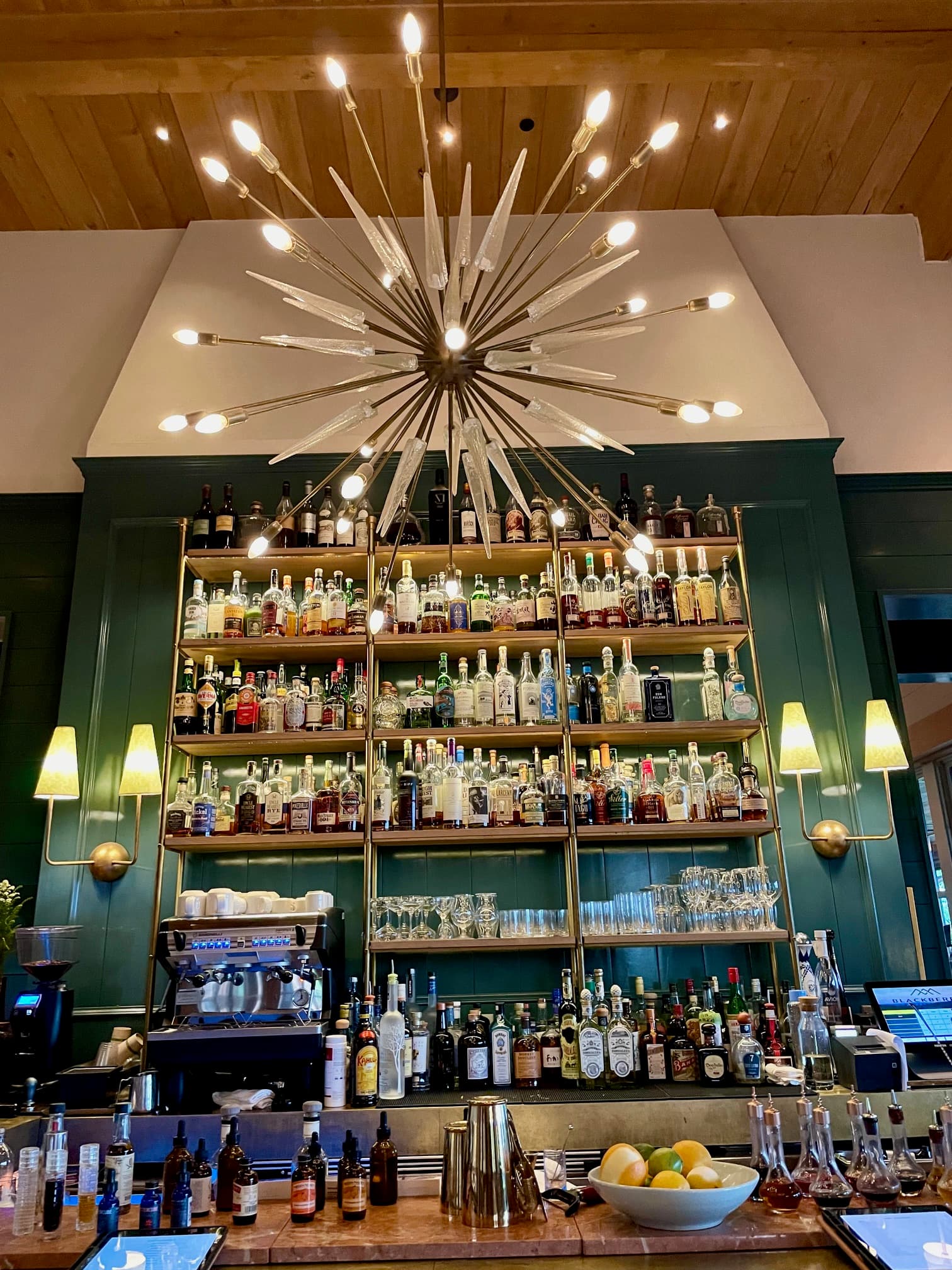 A restaurant bar with a large mid-century modern chandelier, green walls, sconces and shelves full of liquor.