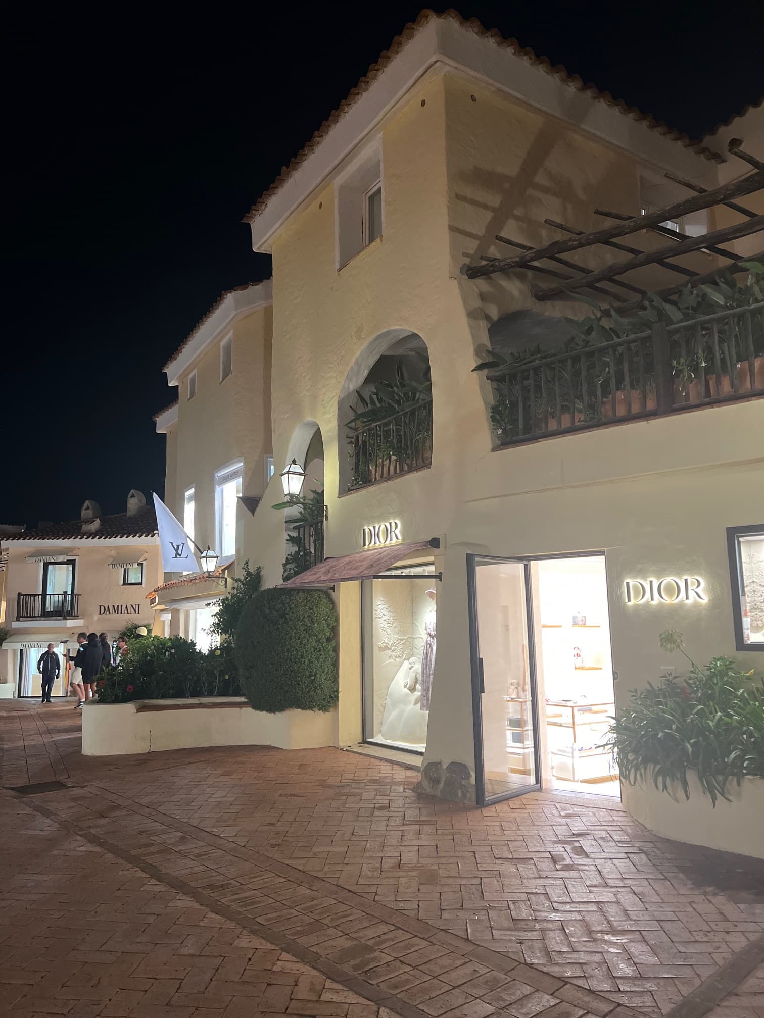 A view of the hotel exterior, complete with a brick road, open door lit from the inside, bushes and a balcony on the top, second level.
