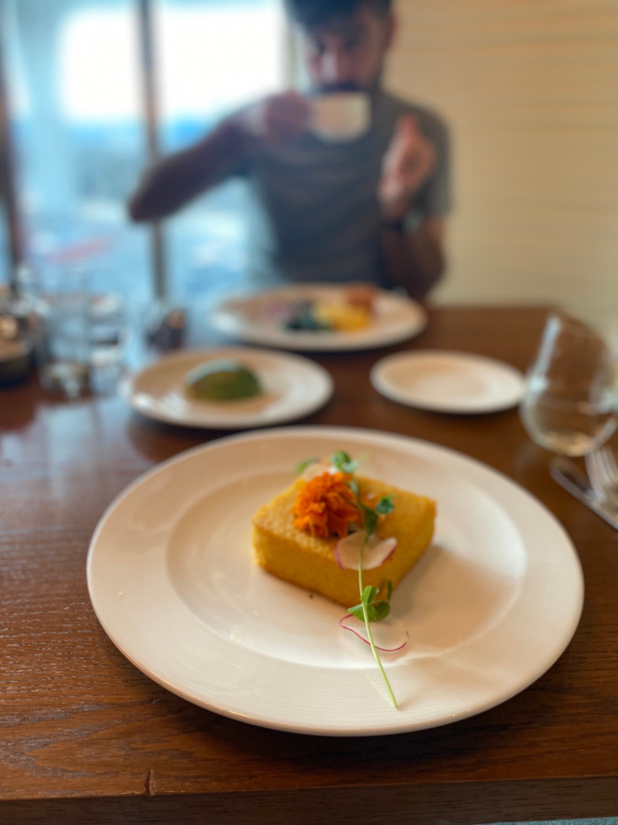 A view of a yellow pastry on top of a white plate. There is a person sipping coffee out of focus in the background.