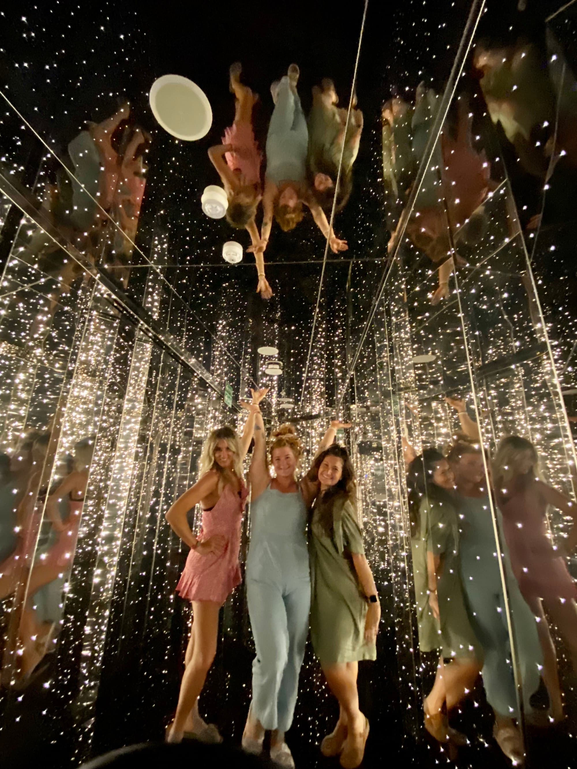 Three women posing beneath reflective mirrors and sparkling lights before a night out.