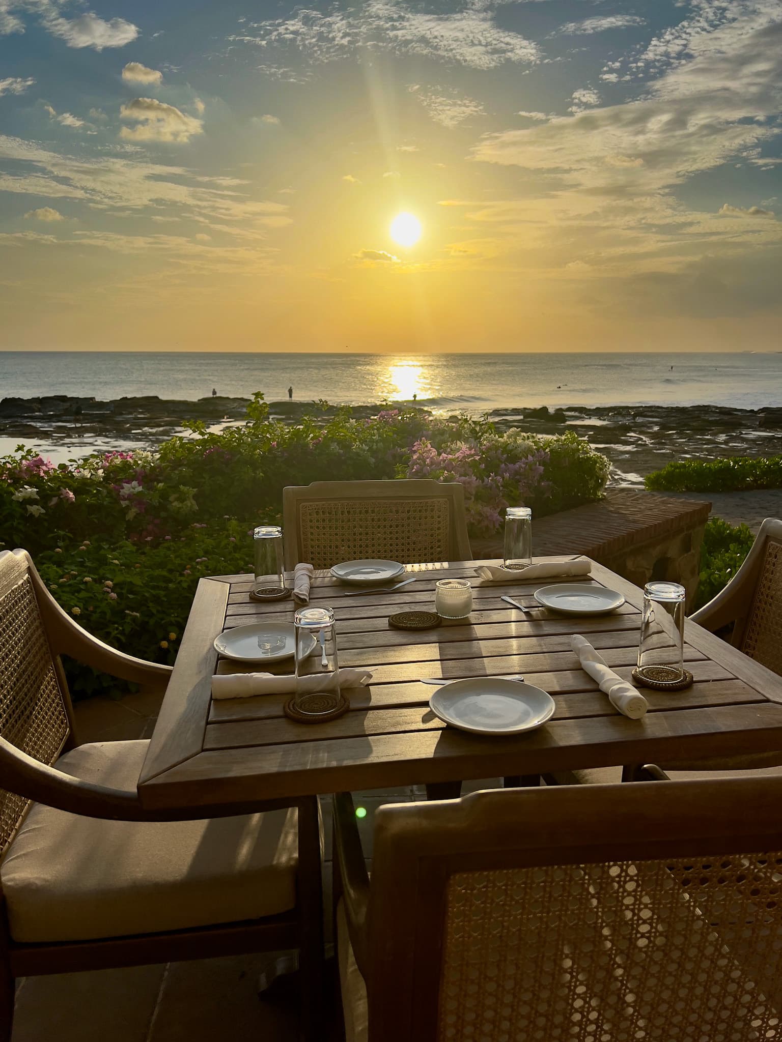 An outdoor dining area with a wooden table, set with plates, glasses and cutlery, overlooking the landscape and sea at sunset.