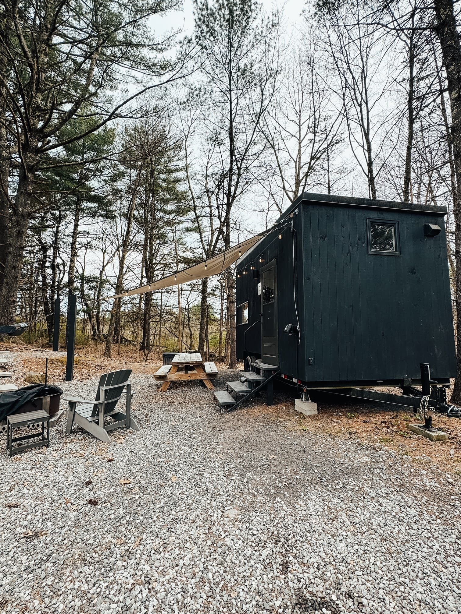 An exterior view of the trailer-style cabin of Getaway House, with an awning, picnic table, chairs, and a fire pit on a cloudy day.