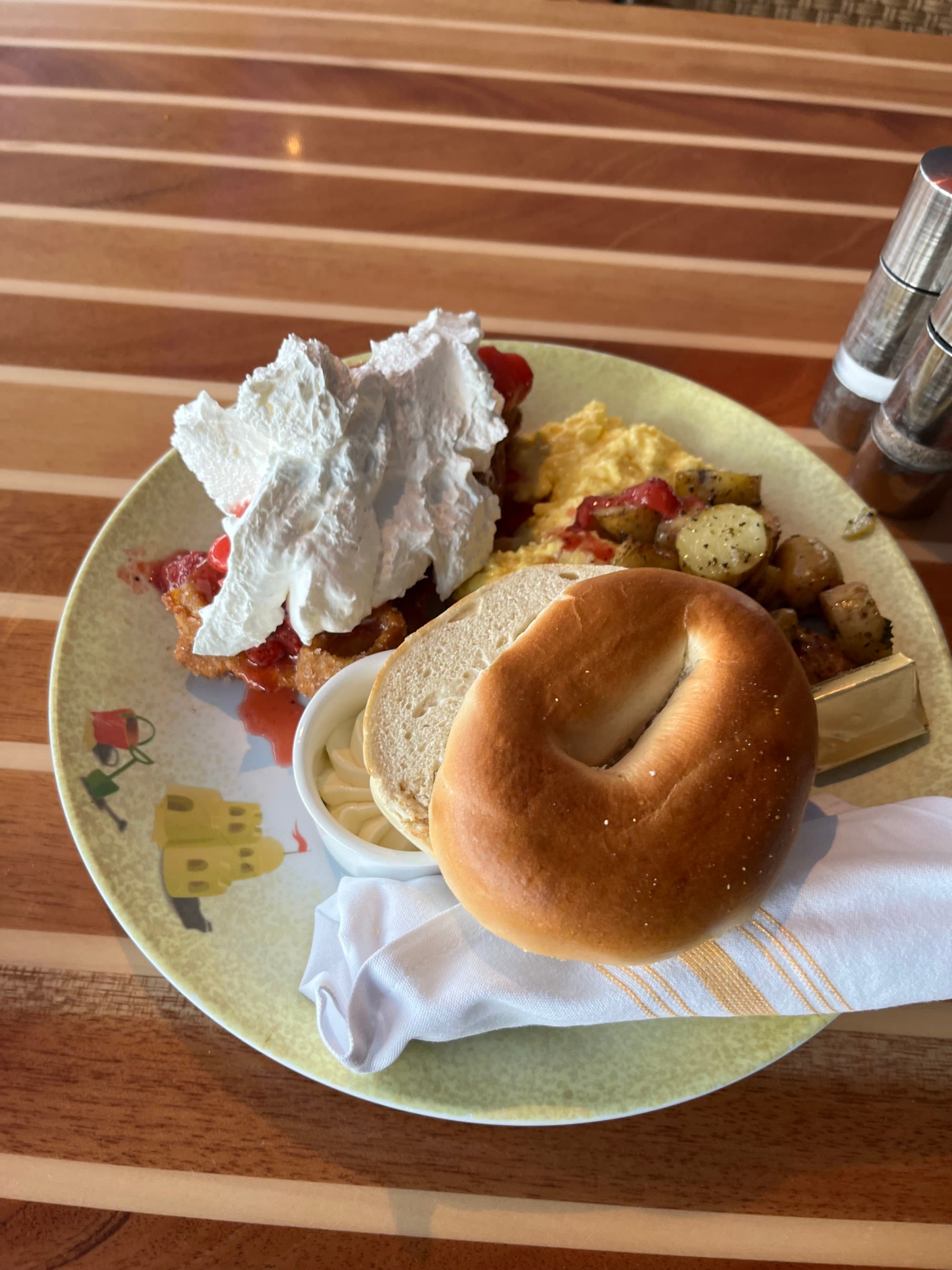 A plate of breakfast foods, including a bagel, eggs, home fries and waffles covered in strawberries and whipped cream.
